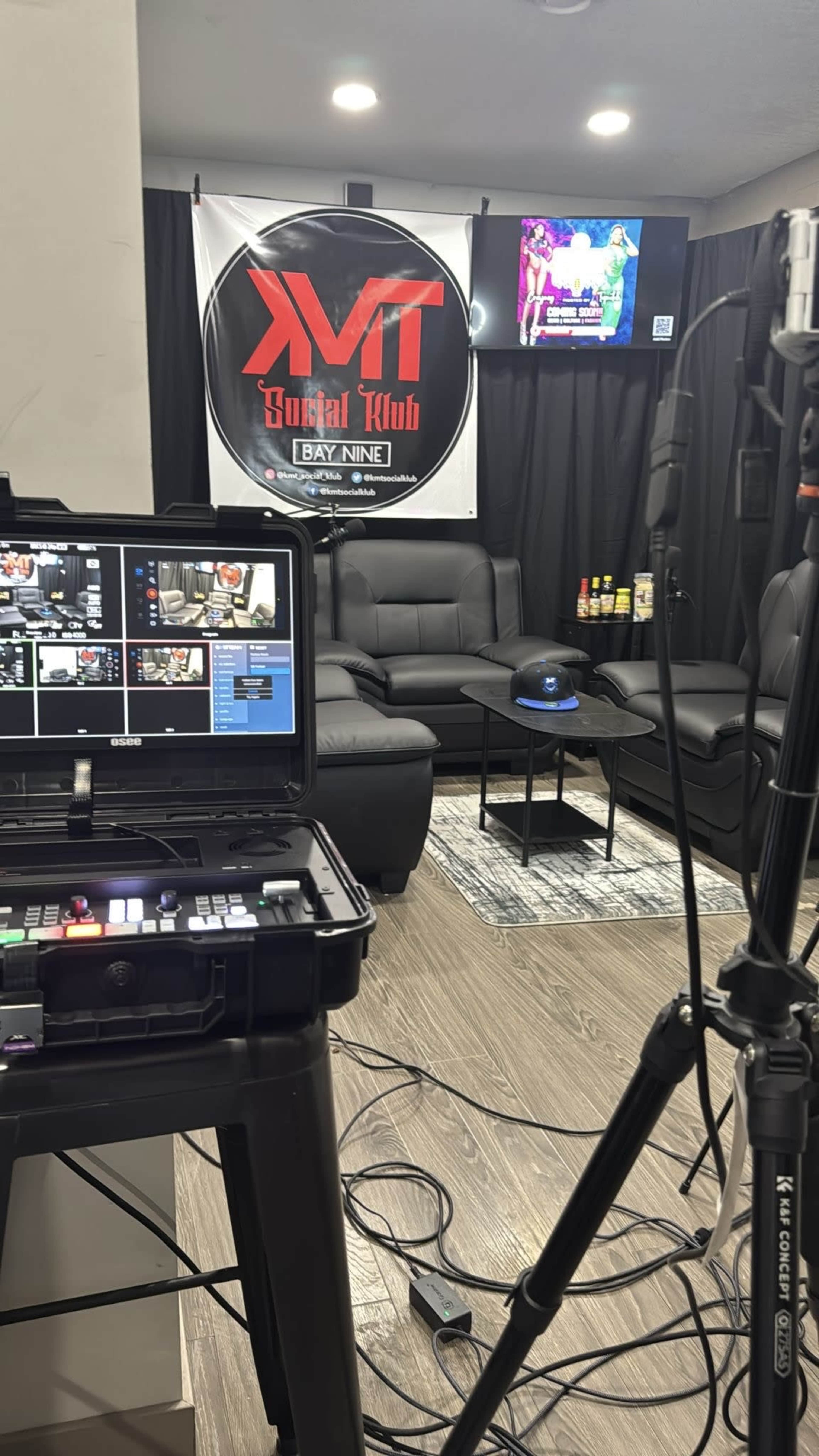 The image shows a production setup for a video recording or broadcast, featuring a black couch, a TV mounted on the wall, and a control panel in the foreground.