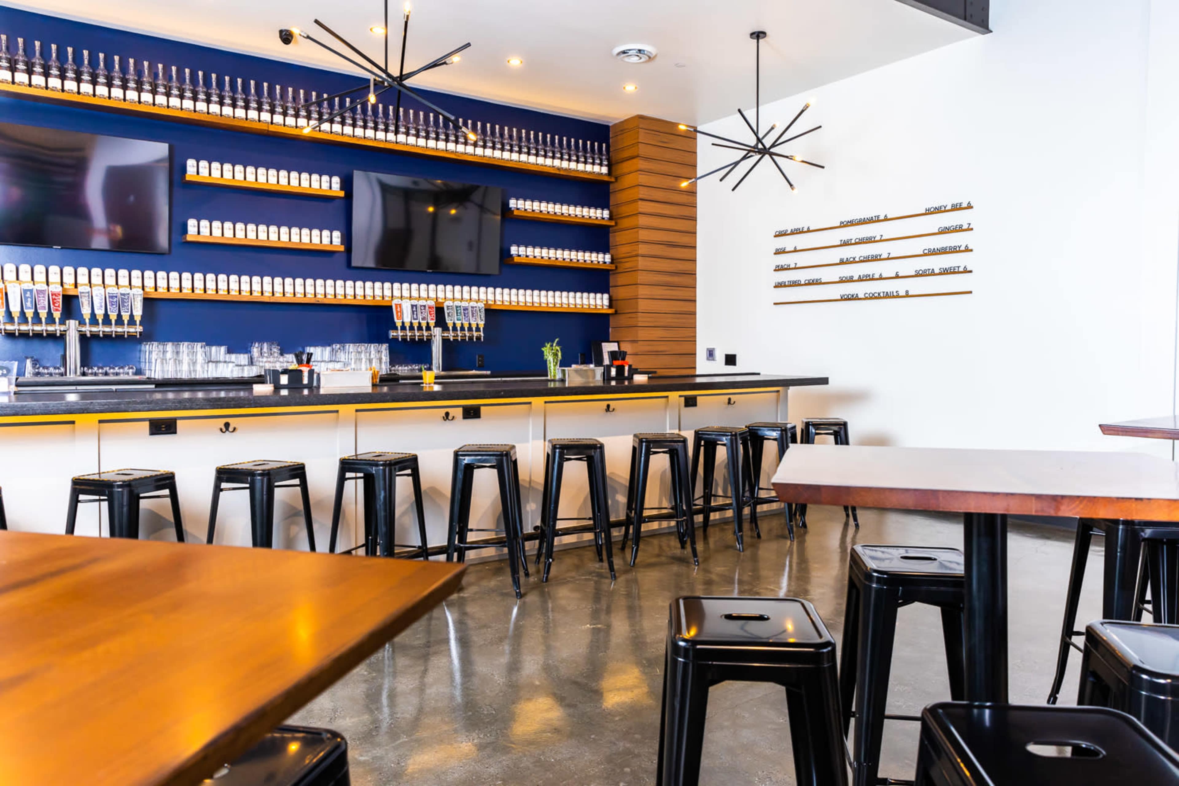 The image shows a modern bar area with a long counter, metal stools, and a wall lined with beverages.