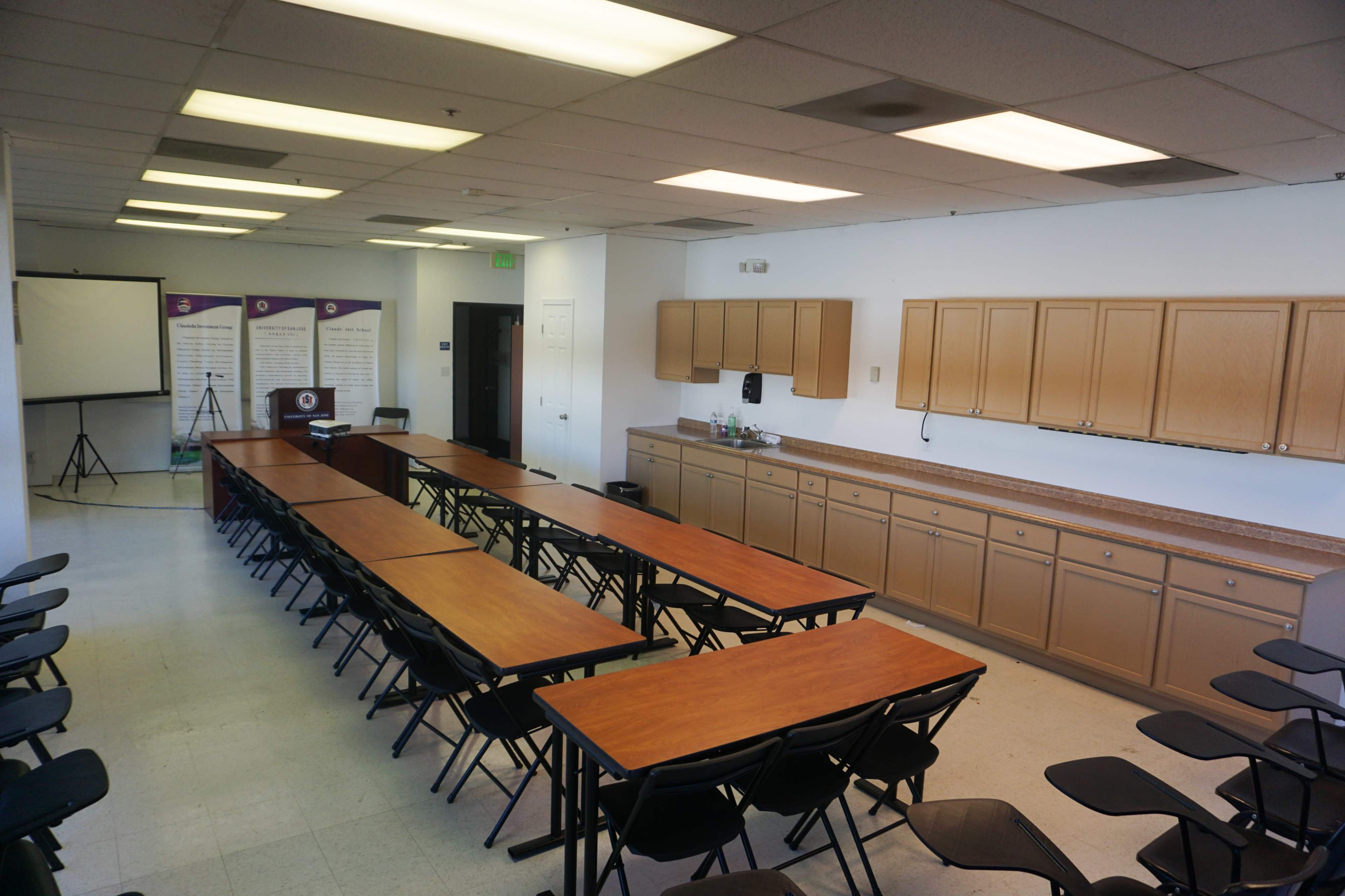 A meeting room features several rows of folding tables and chairs, with a kitchenette along one wall and a projector screen set up in the corner.