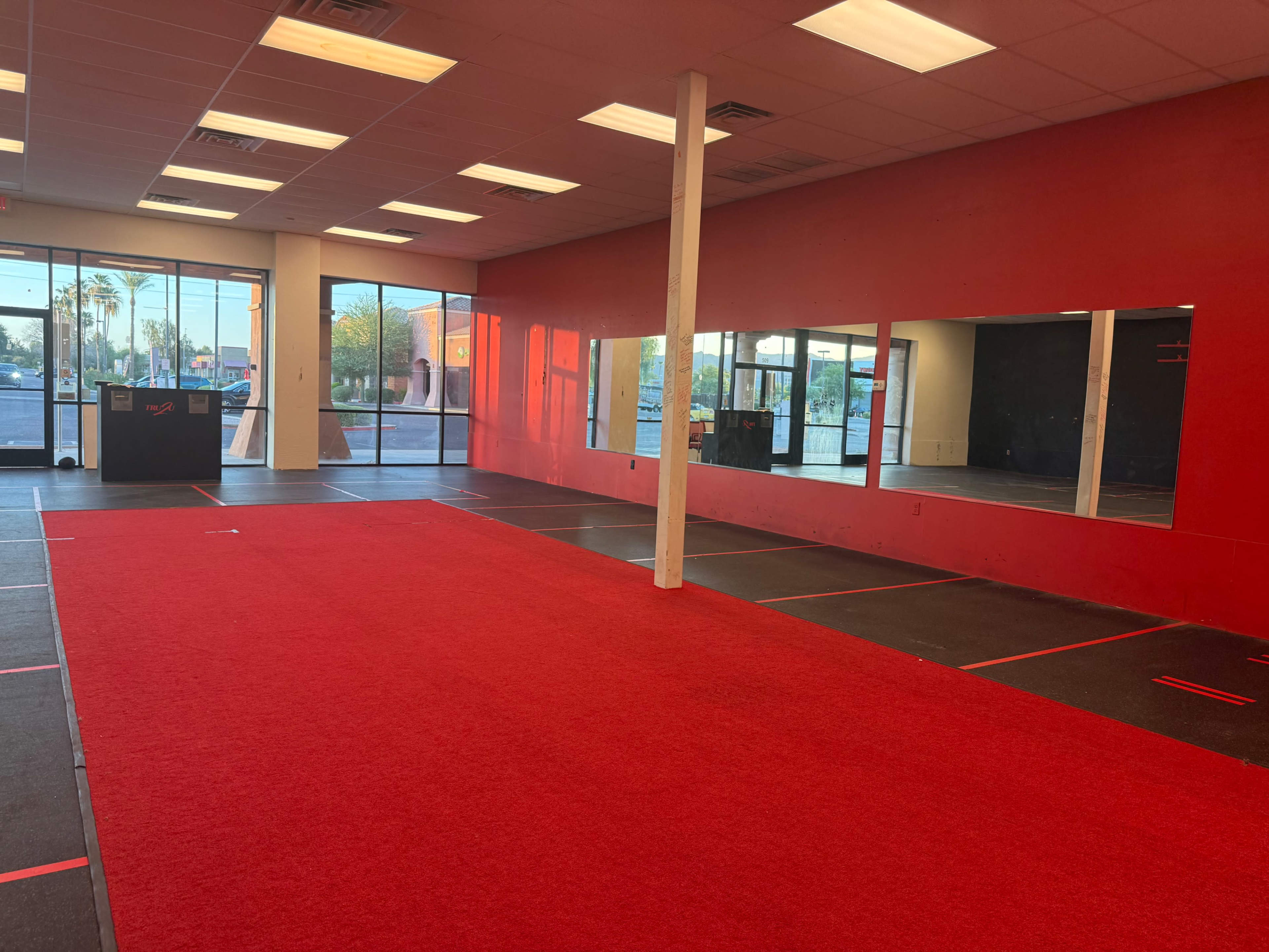 The image shows an empty training space with red carpeting and black flooring, featuring large mirrors on one wall and windows that allow natural light to enter.