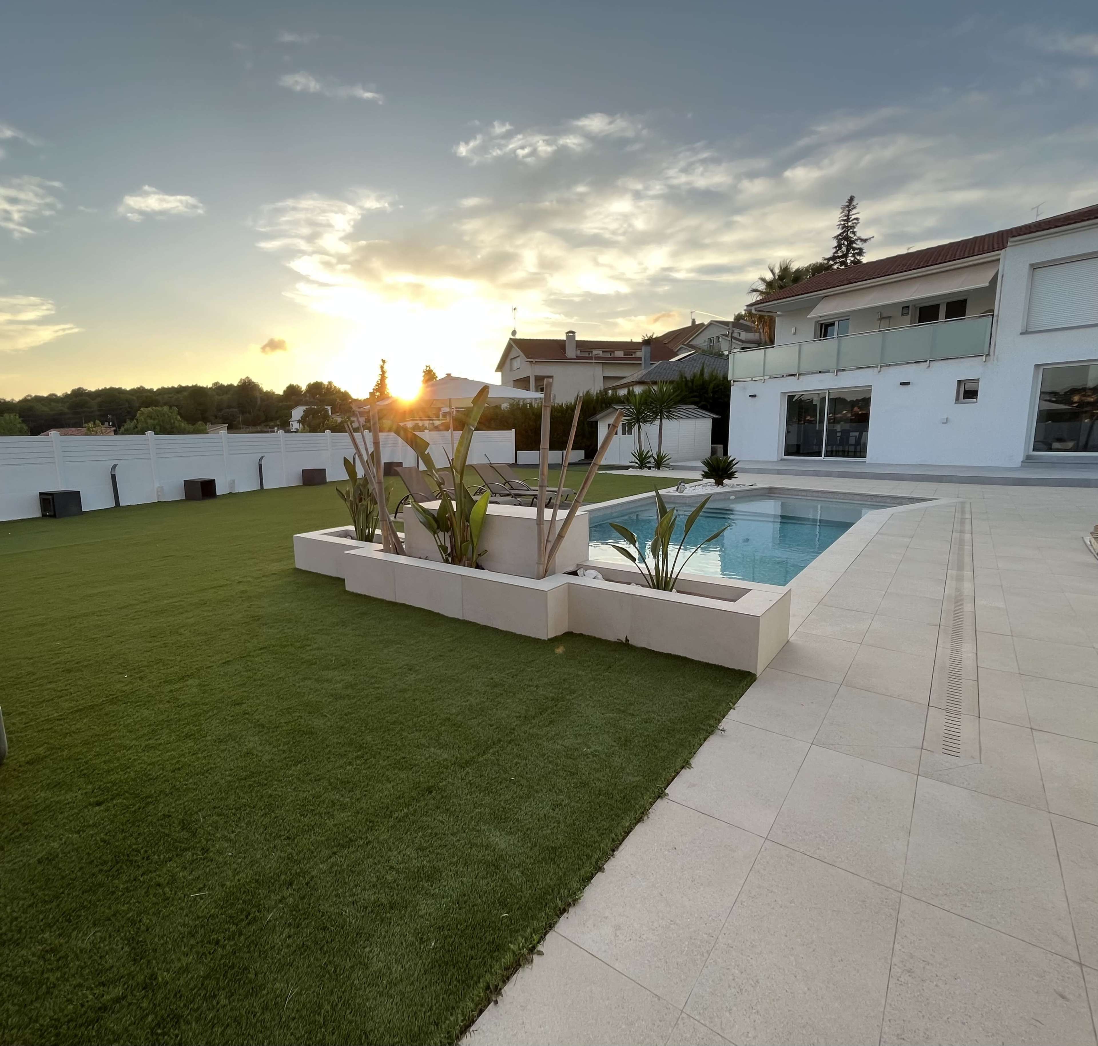 A modern backyard with a swimming pool, landscaped plants, and a sunset in the background.