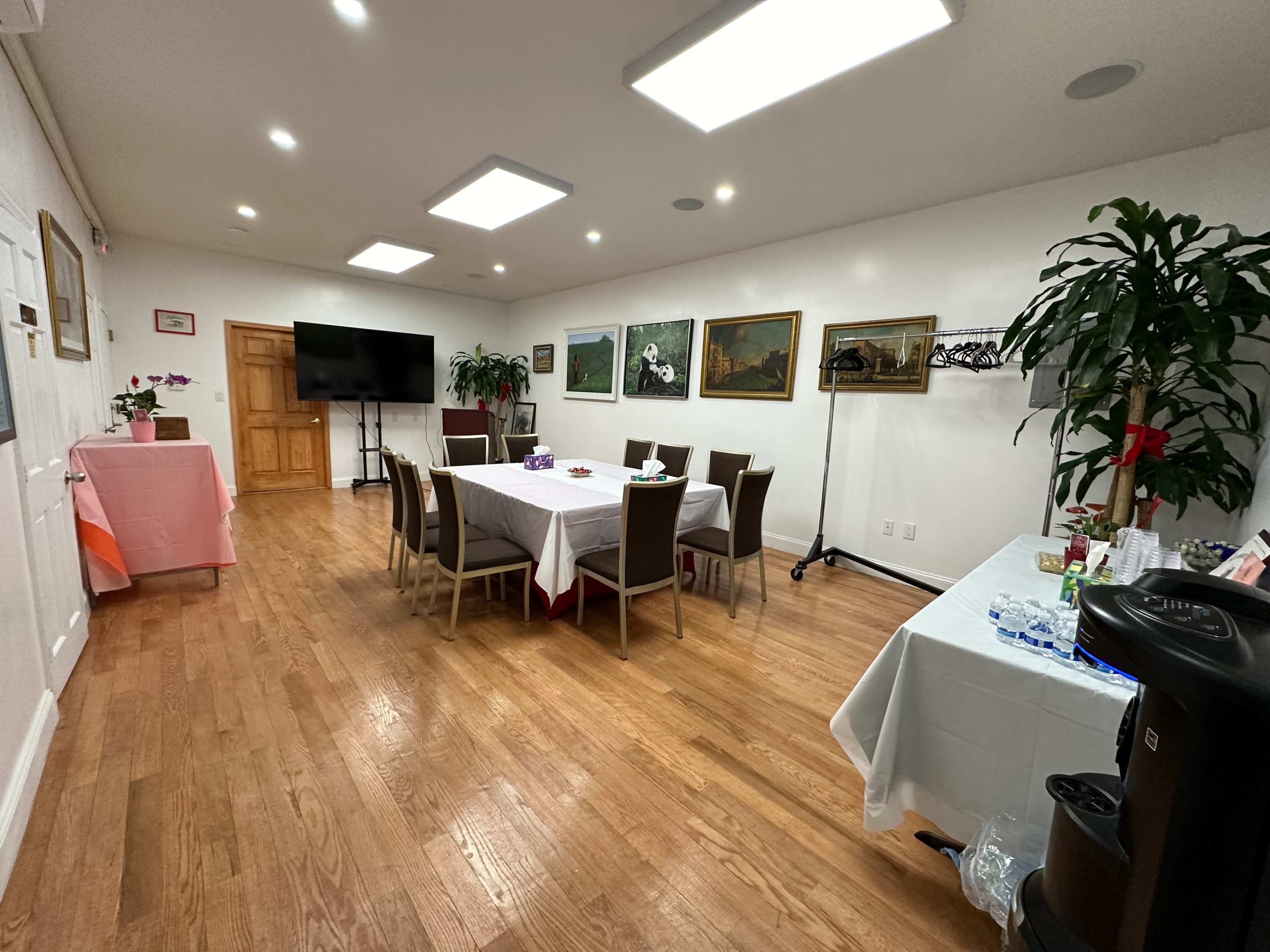 The image shows a meeting room with a large table set for an event, surrounded by chairs, and displays paintings on the walls, with refreshments on a side table.
