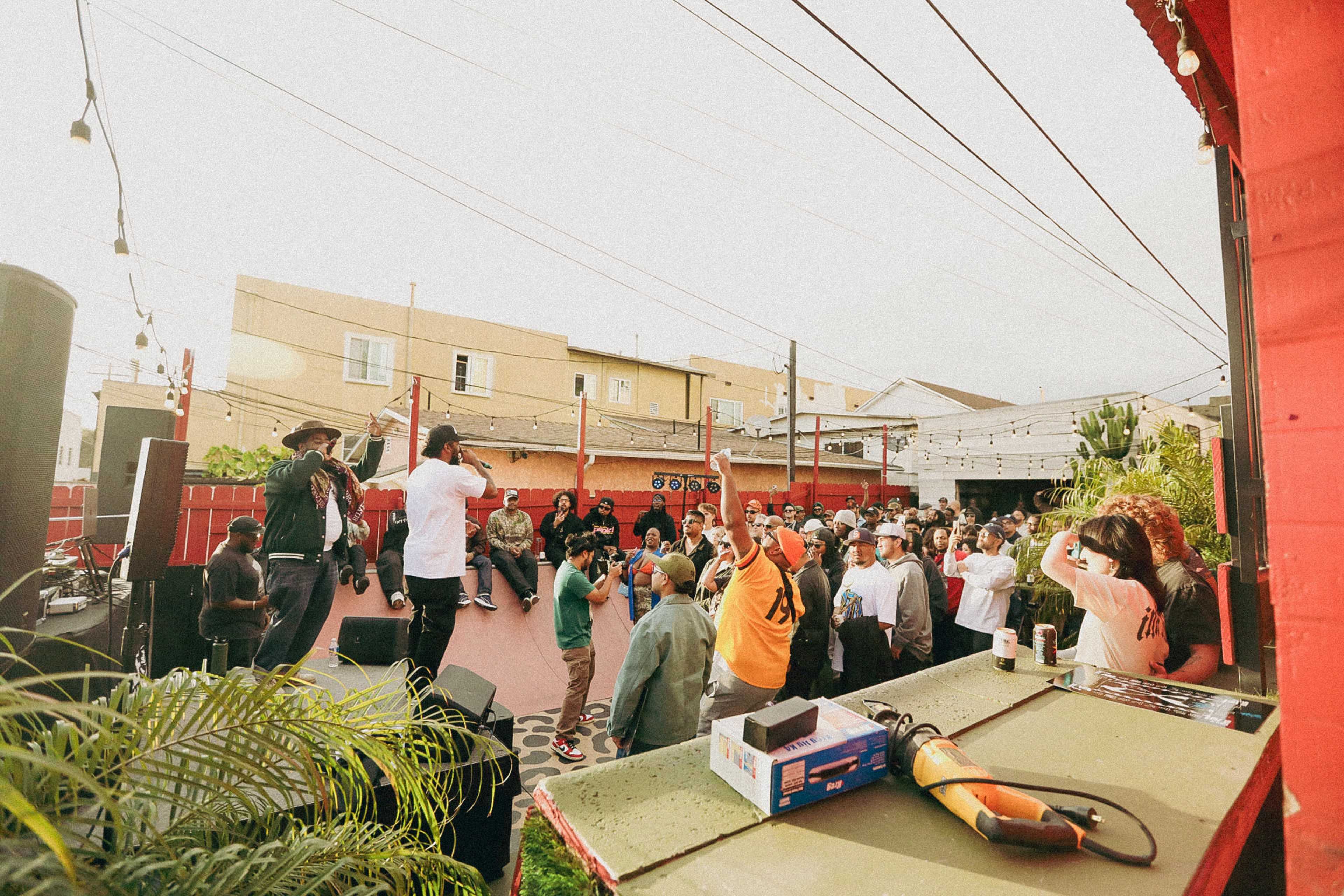 A crowd gathers in an outdoor space with a stage as performers engage the audience amidst a backdrop of colorful buildings.