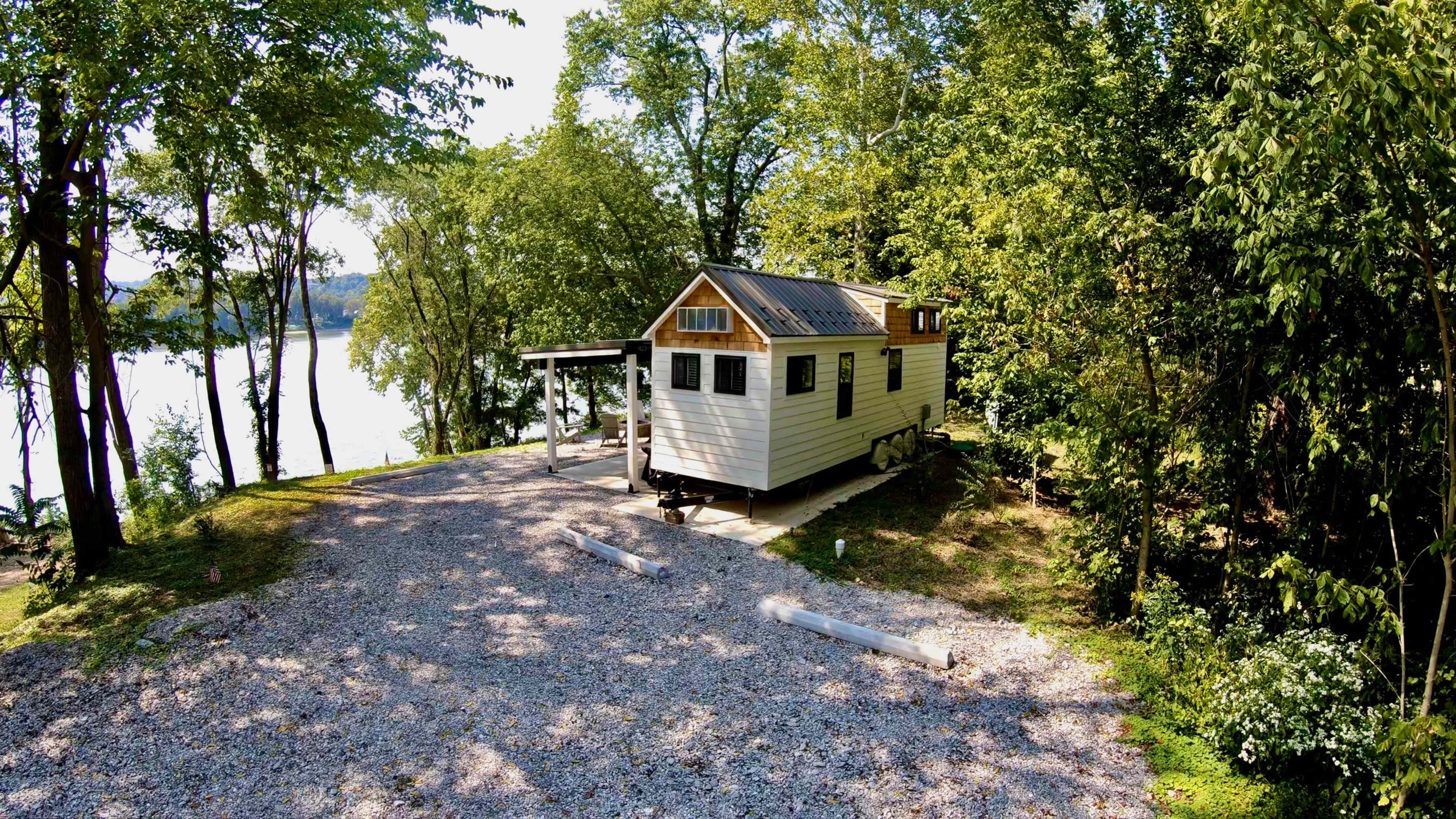 A small cabin with a sloped roof is parked on a gravel driveway beside a lake, surrounded by trees.