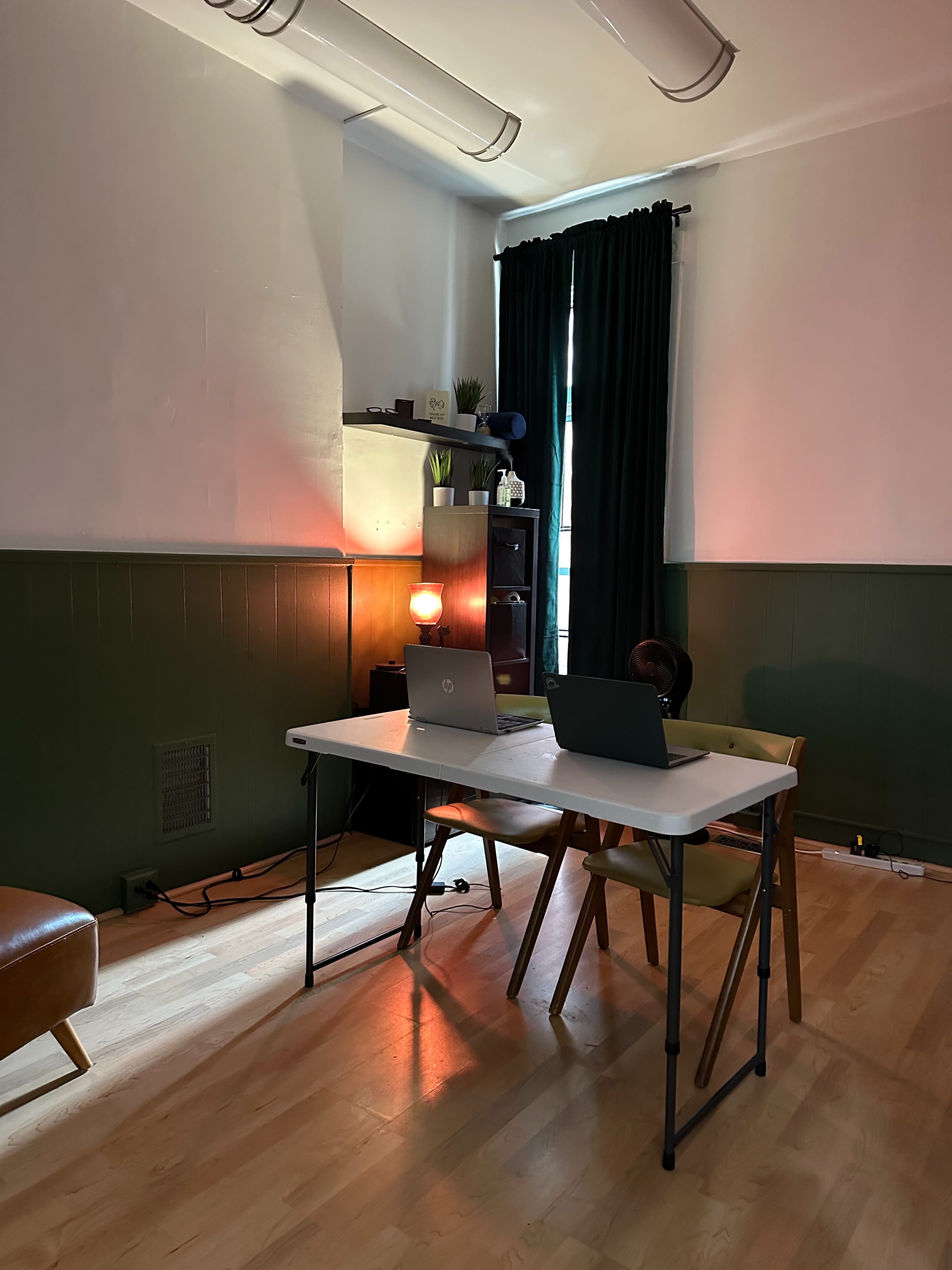 A simple workspace is set up in a room with a table, two laptops, a chair, and dark green walls.