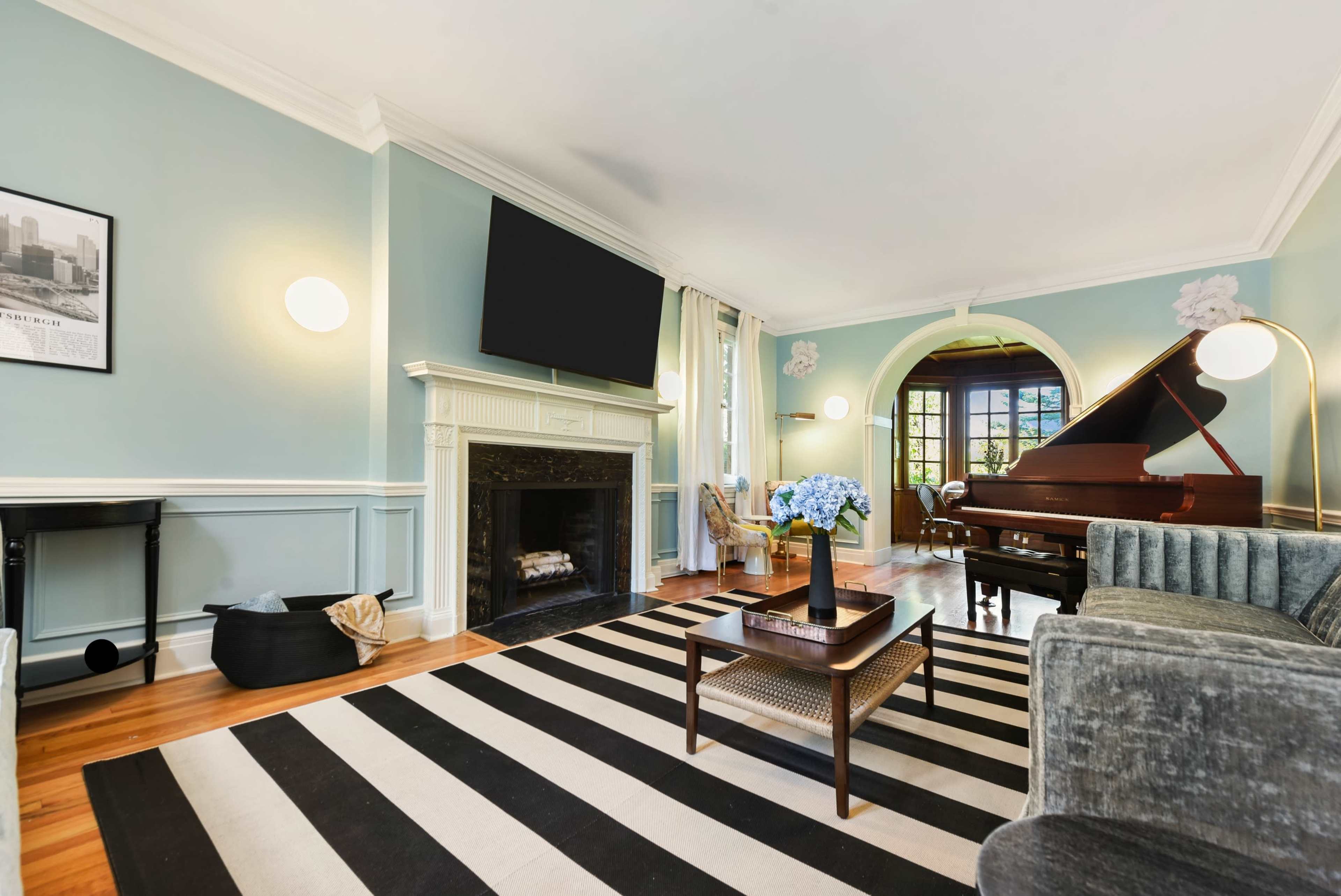 The image shows a living room with a fireplace, a grand piano, and a striped rug, featuring light blue walls and natural light coming in from a large window.