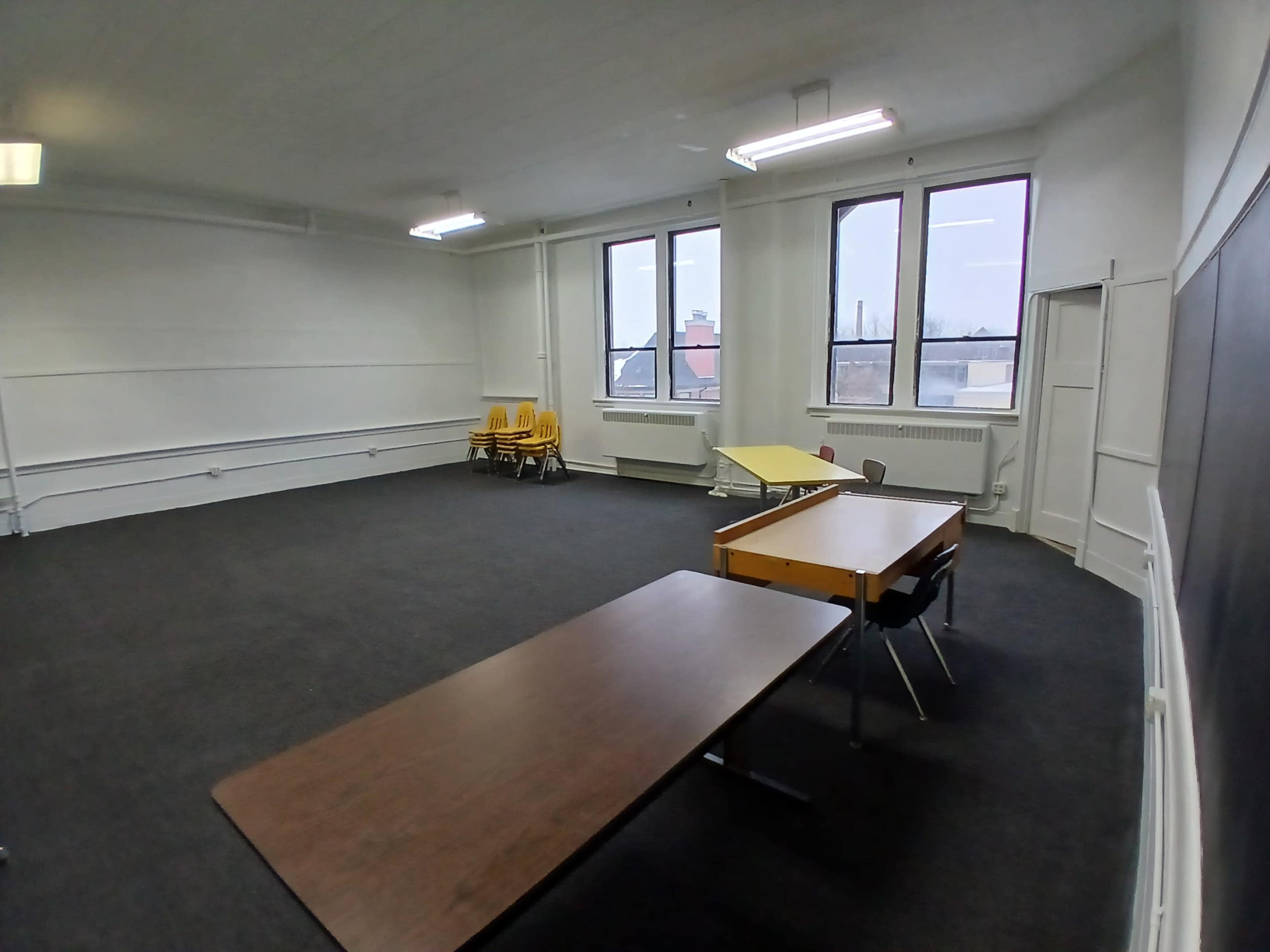 The image shows a spacious, empty classroom with a few stacked chairs and desks placed in various positions.