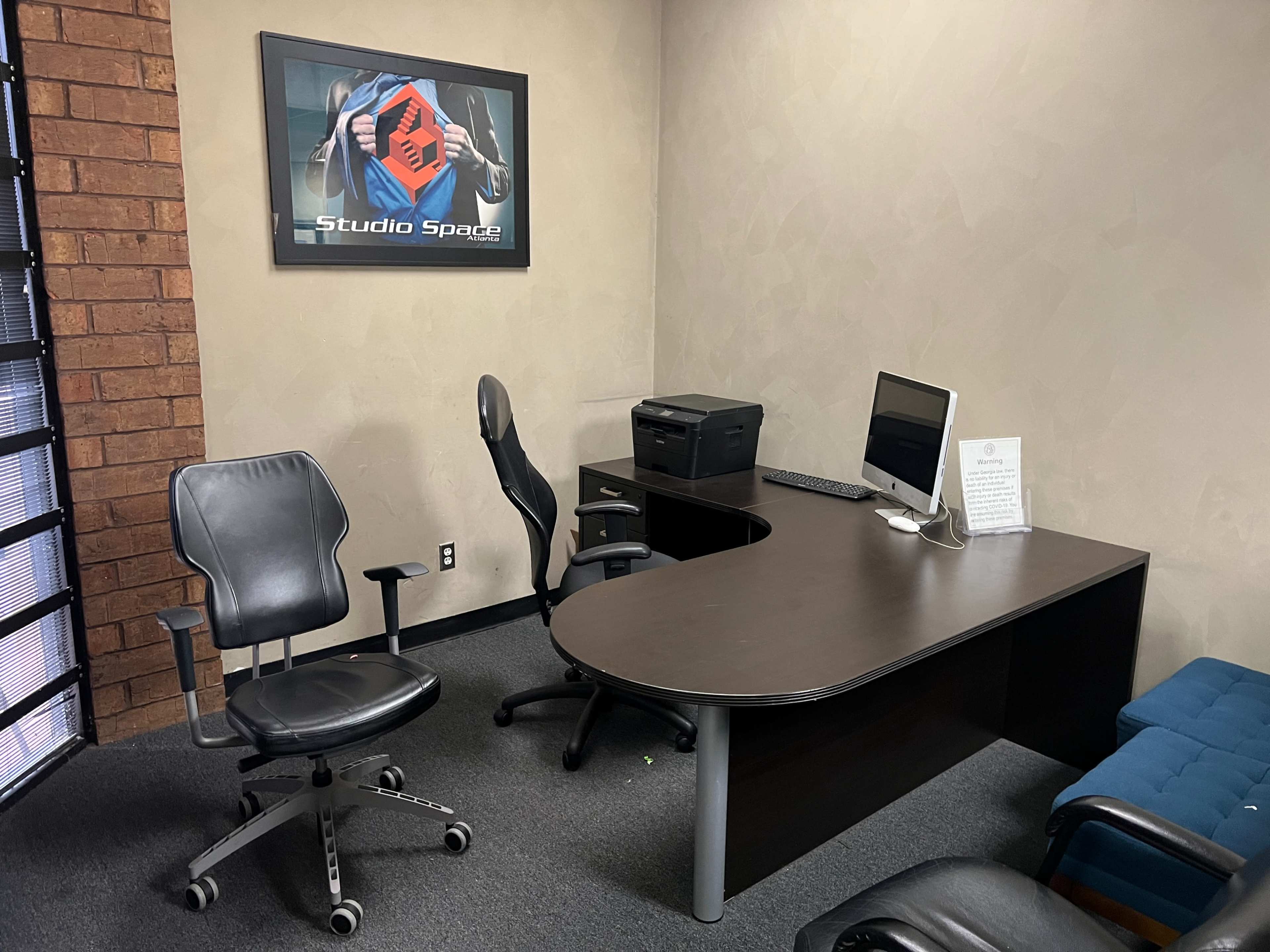 The image shows a small office with a brown desk, a computer monitor, a printer, and two chairs, one of which is a rolling office chair.