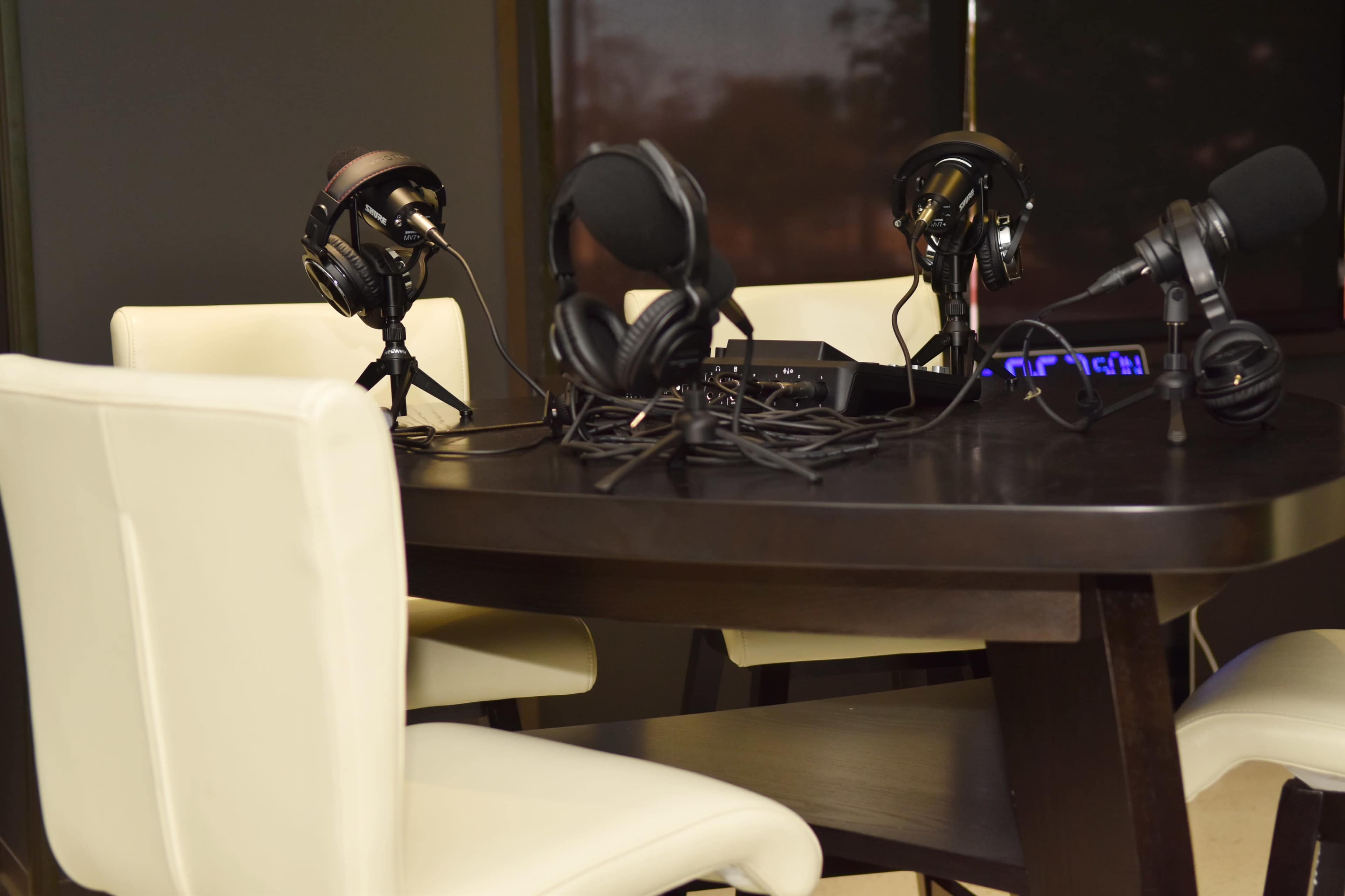 A wooden table is set with three microphones and headphones, surrounded by two white chairs.