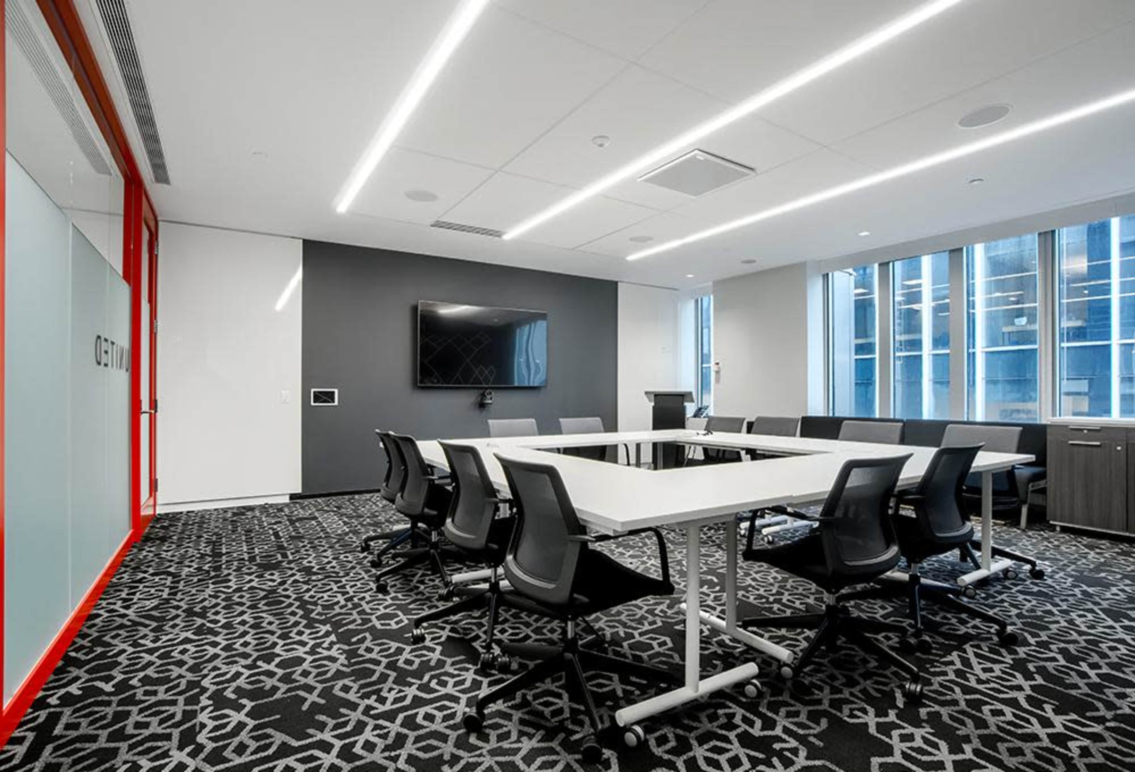 A modern conference room featuring a large rectangular table surrounded by chairs, with a television mounted on the wall and large windows providing natural light.