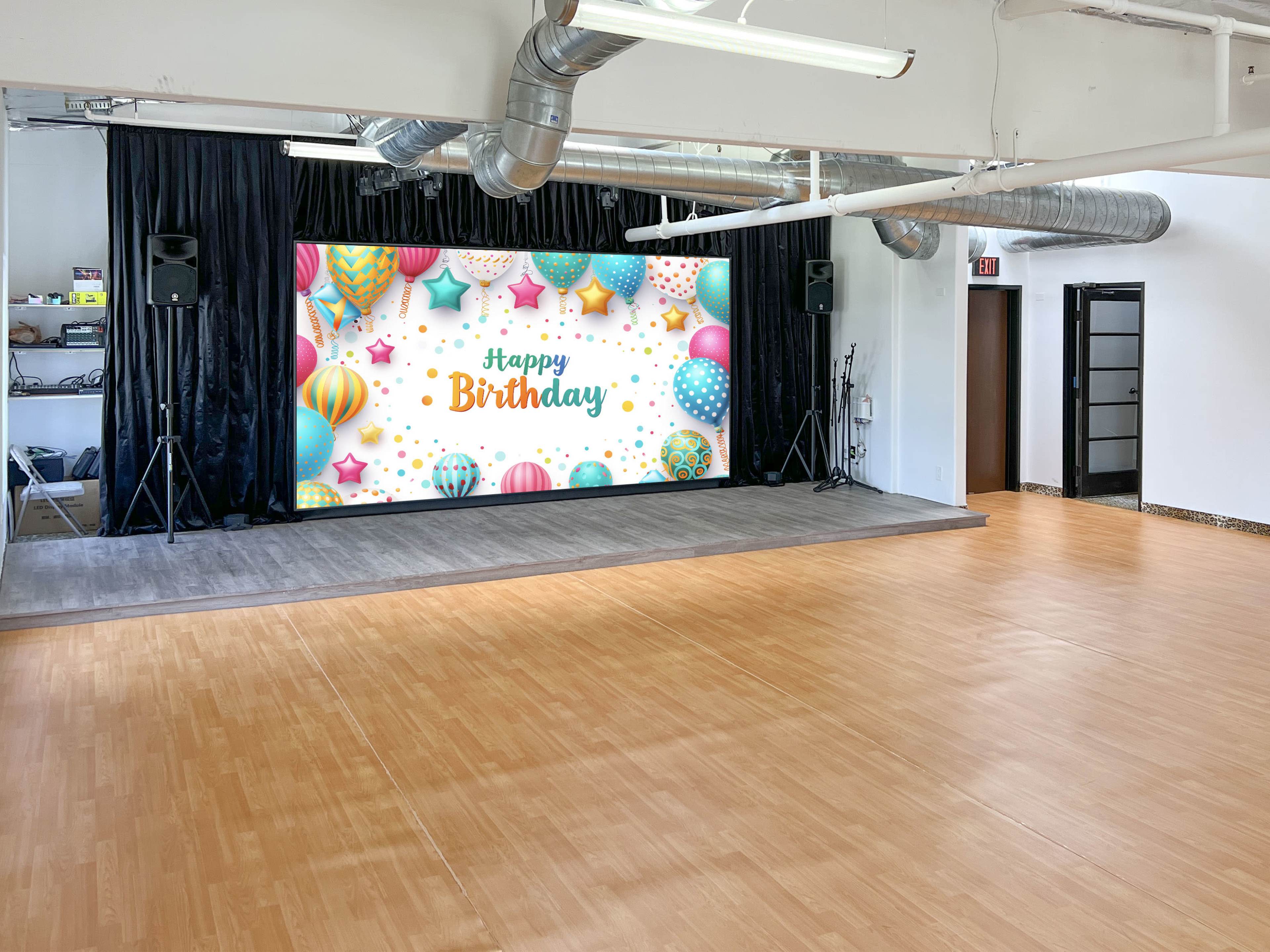 The image shows a spacious room with a large birthday-themed backdrop featuring colorful balloons and the text "Happy Birthday," accompanied by speakers on either side.