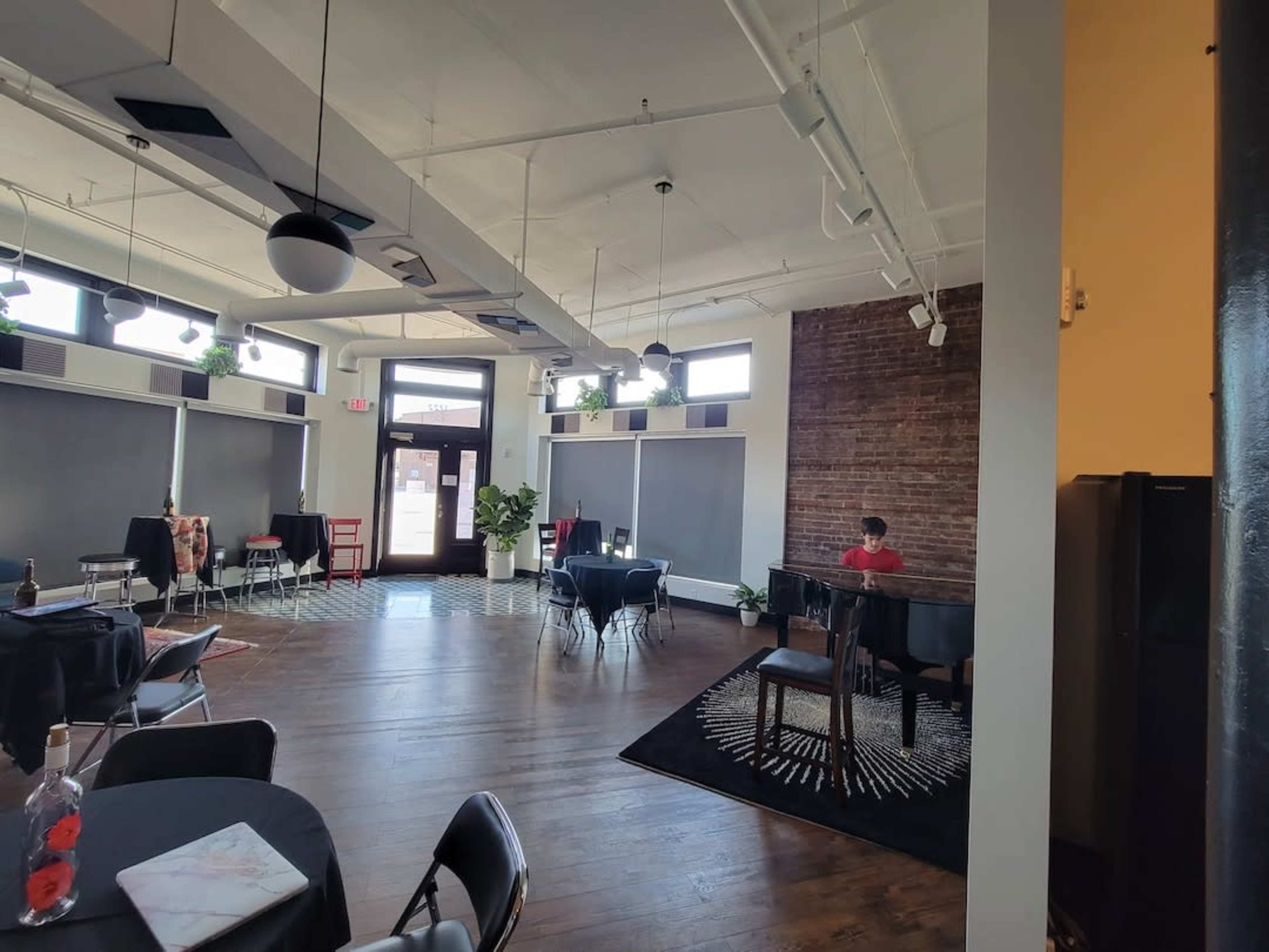 A spacious interior features wooden floors, tables set for dining, and a person seated at a piano against a backdrop of exposed brick.