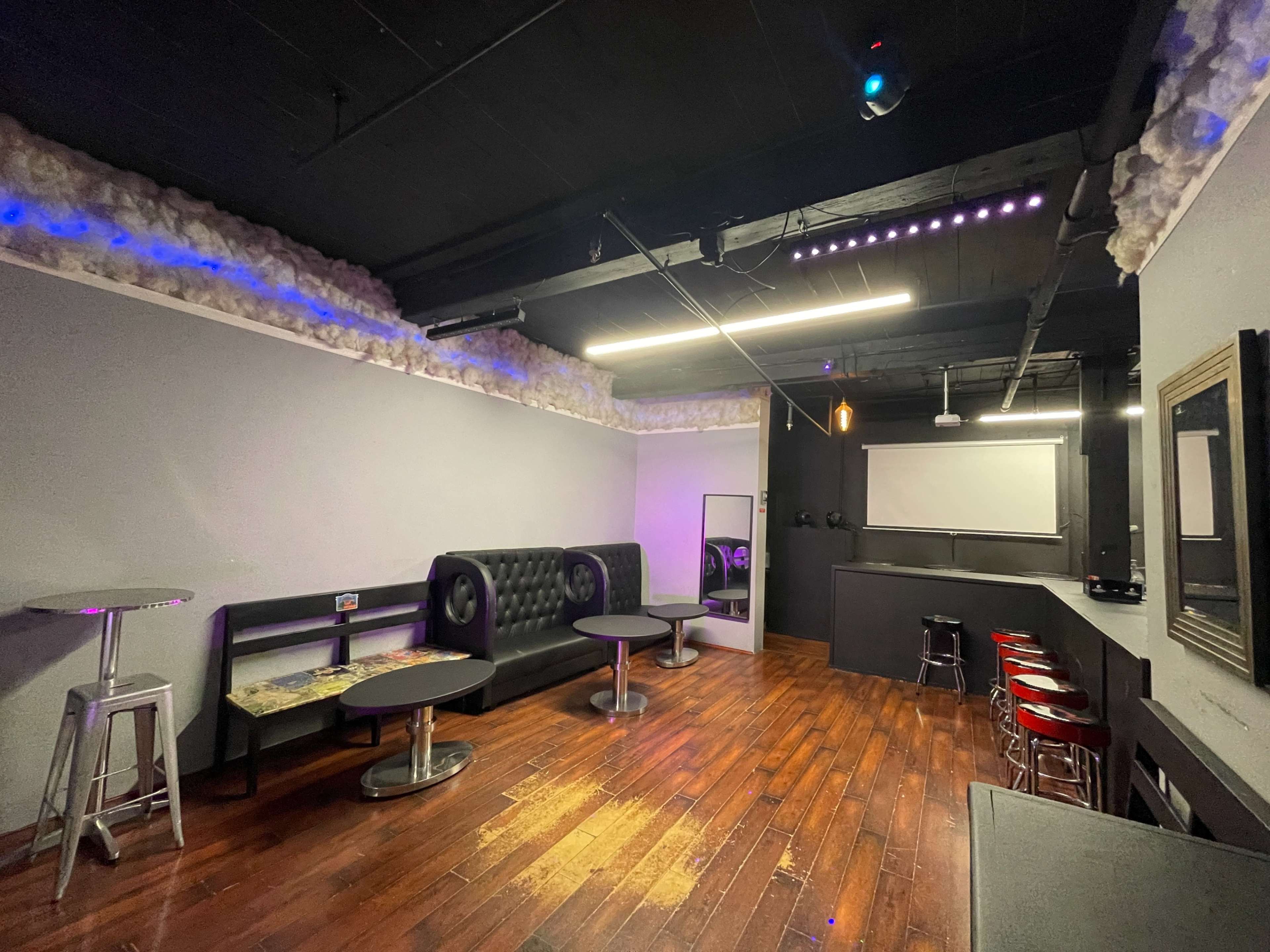 A dimly lit lounge area with black leather furniture, wooden flooring, and a projection screen on the wall.