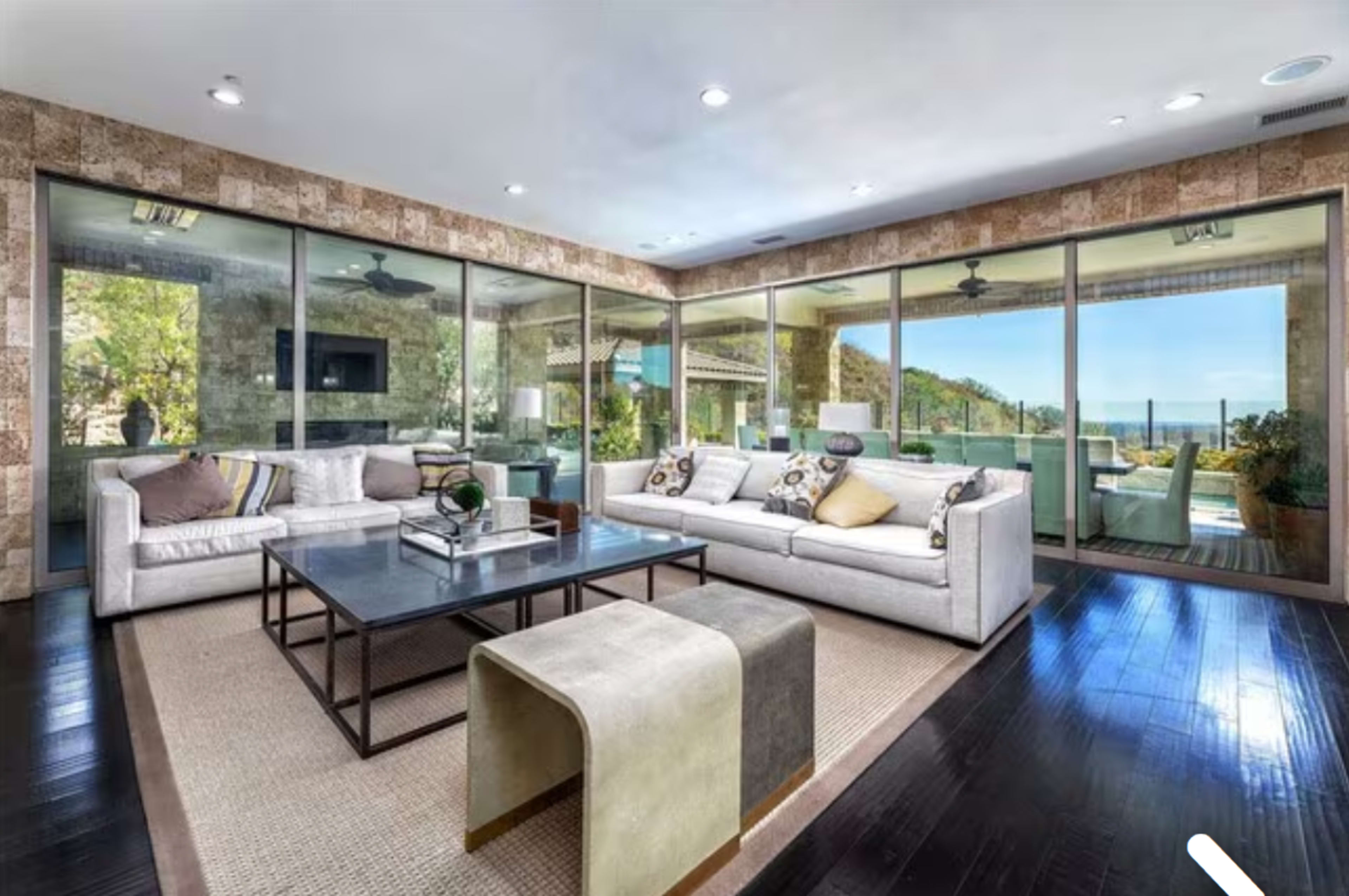 The image shows a modern living room featuring large glass windows that provide a view of the outdoors, with a seating area that includes a light-colored sofa and a central coffee table.