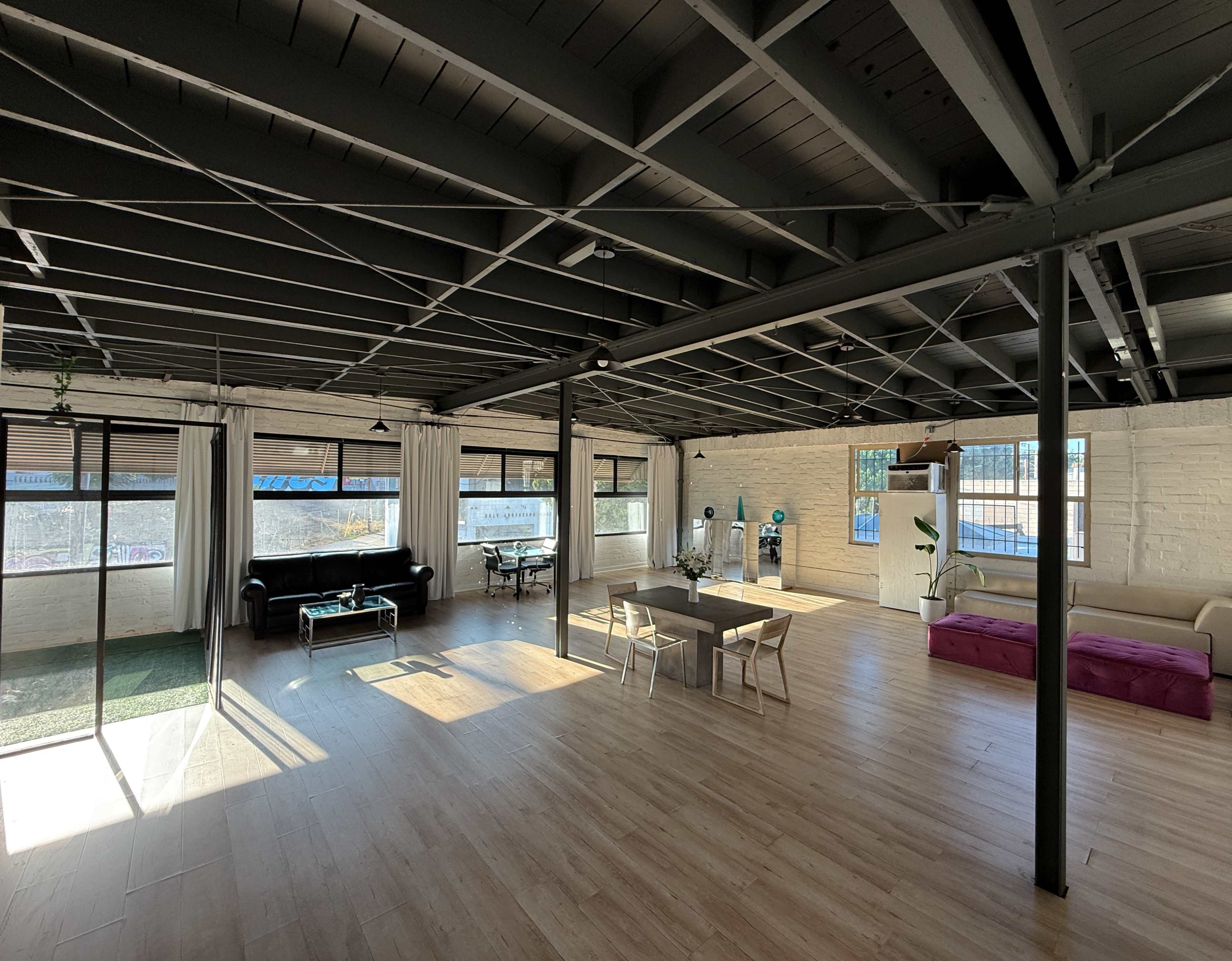 The image shows a spacious, well-lit interior with exposed beams, featuring a mix of seating, tables, and large windows.