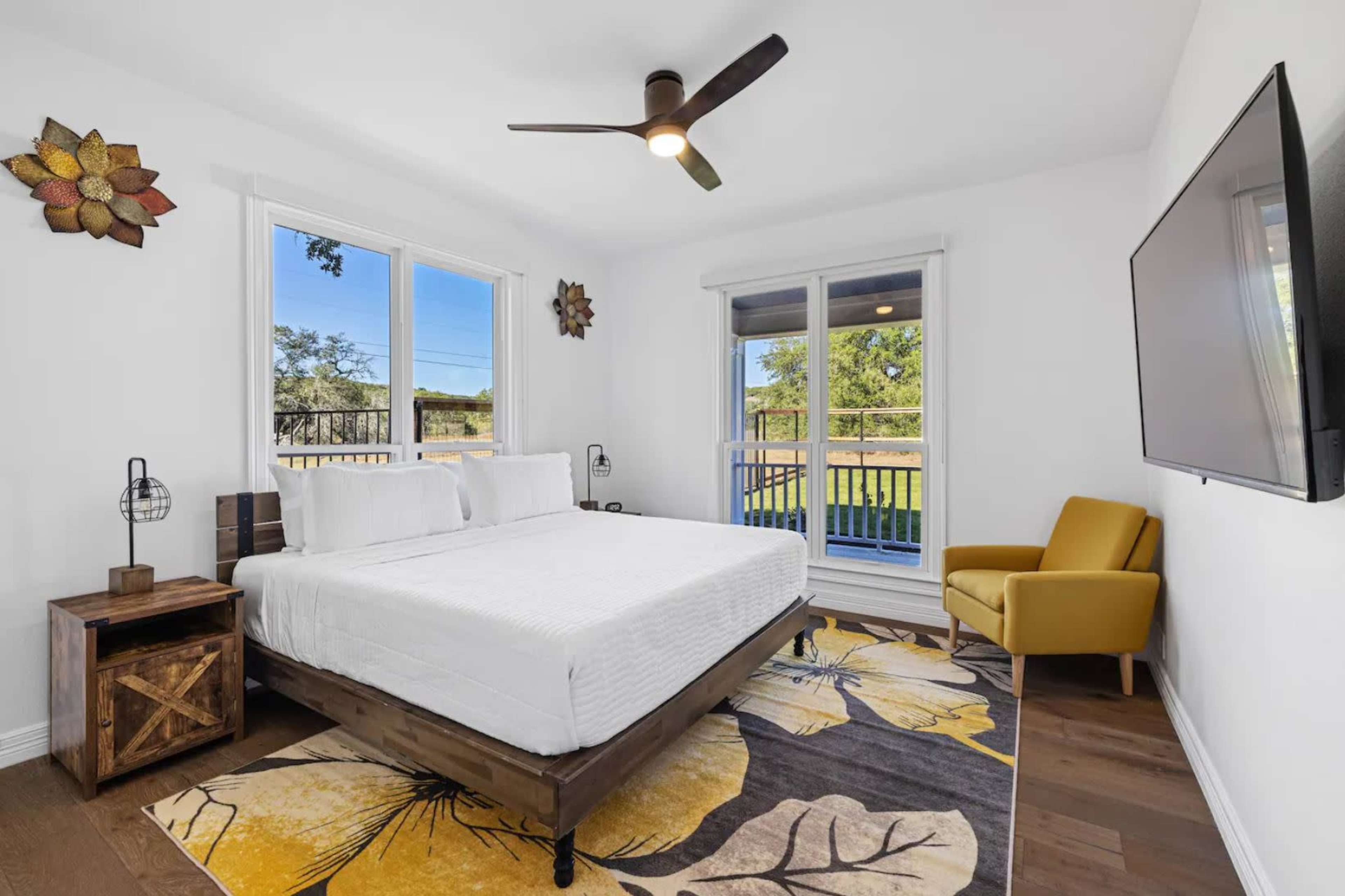 A modern bedroom features a bed with white linens, a yellow armchair, a wooden nightstand, and large windows overlooking a green outdoor area.