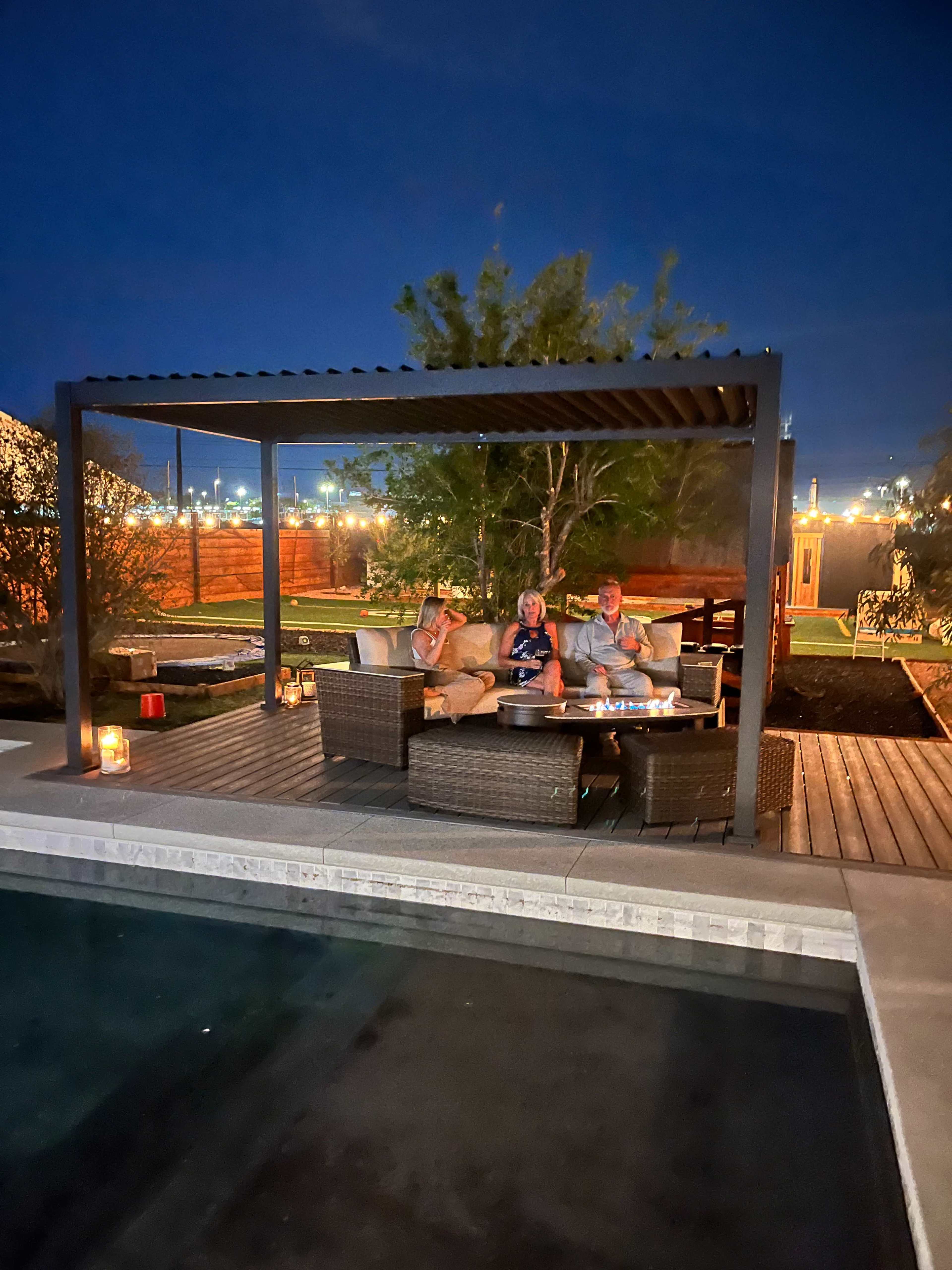 A group of three people sit under a covered patio near a pool, surrounded by lanterns and greenery at night.