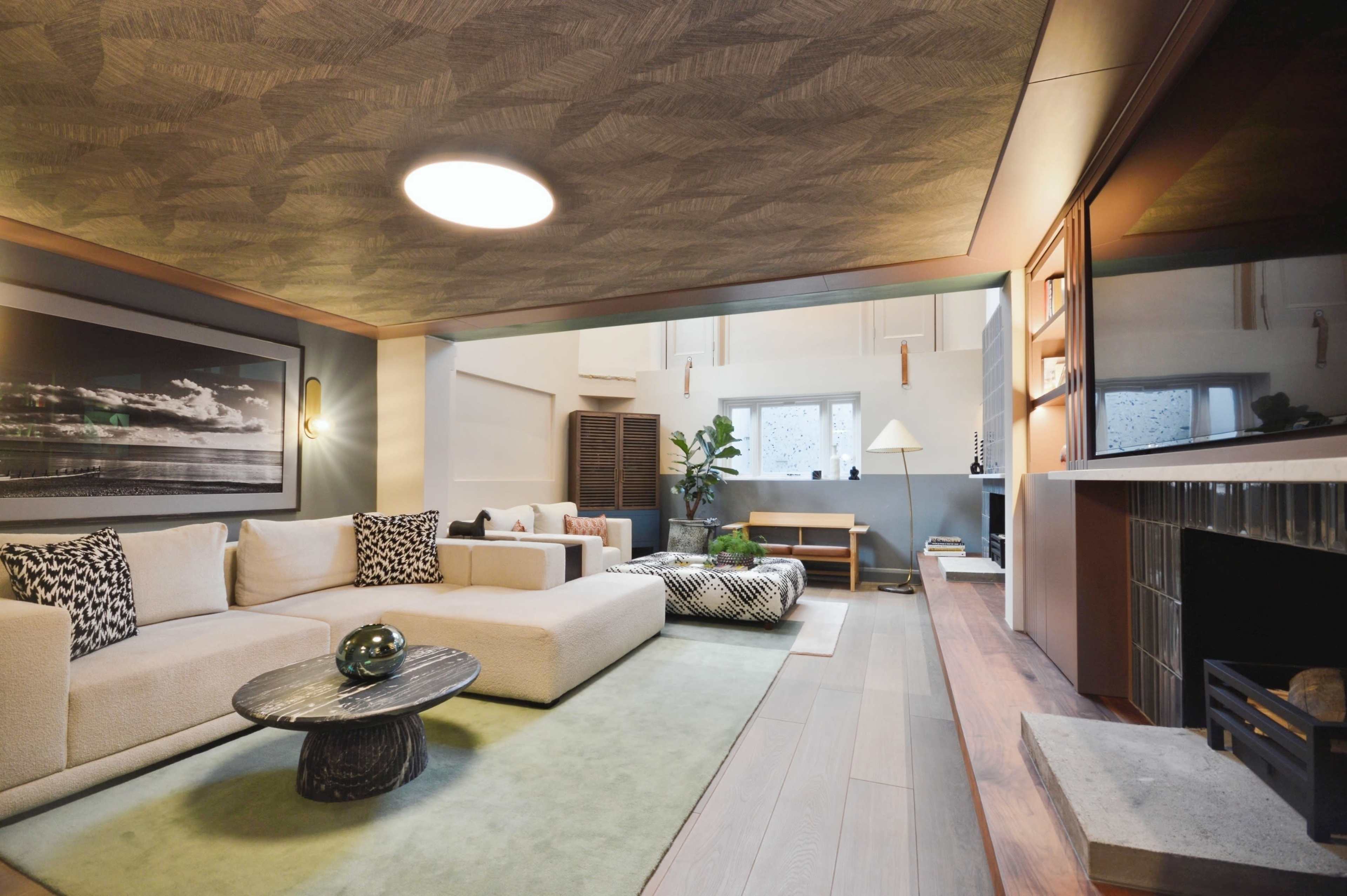 The image shows a modern living room featuring a large beige sectional sofa, a round coffee table, a patterned rug, and a minimalist side table, with ample natural light coming from a window.