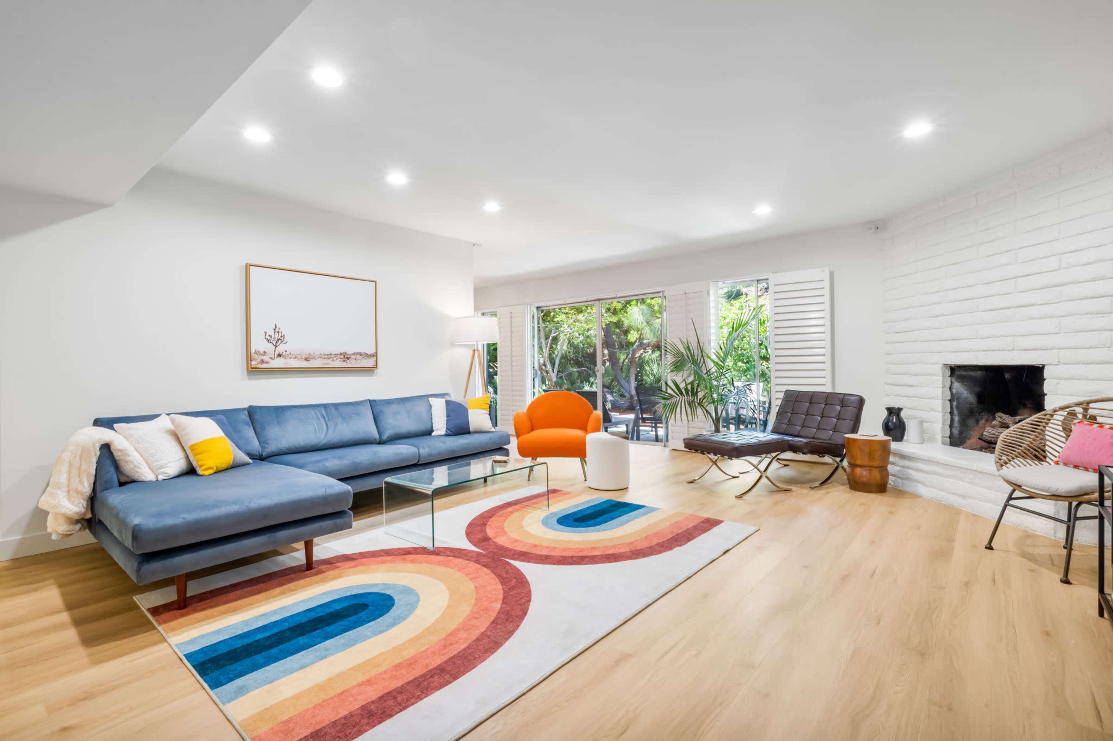 A modern living room with a blue sectional sofa, an orange accent chair, a glass coffee table, and a multicolored area rug, featuring large windows that overlook a green outdoor space.
