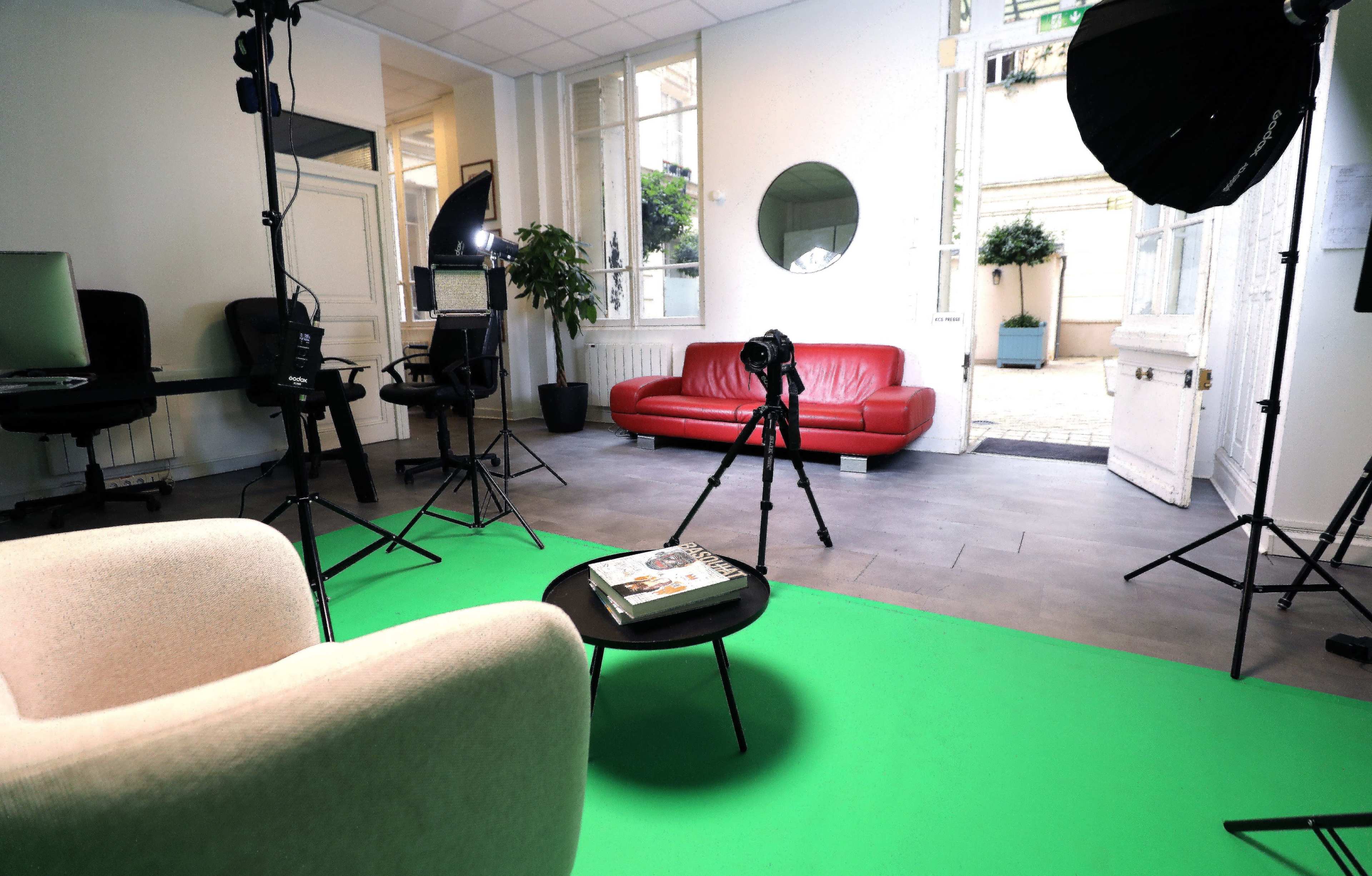 The image shows a studio setup with a green floor, a red couch, a coffee table with a magazine, and camera equipment arranged in a well-lit room featuring large windows.