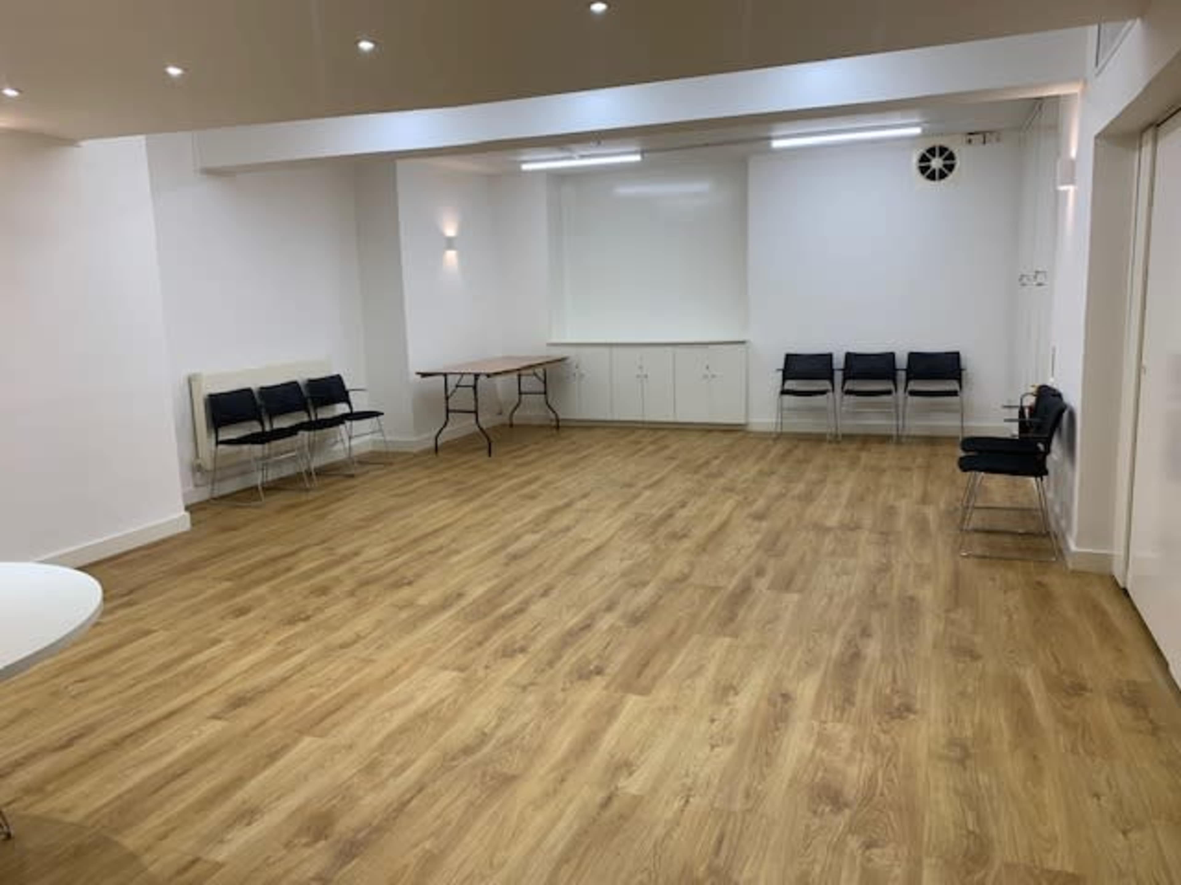 An empty room with wooden flooring, white walls, a table, and several black chairs lined against one side.