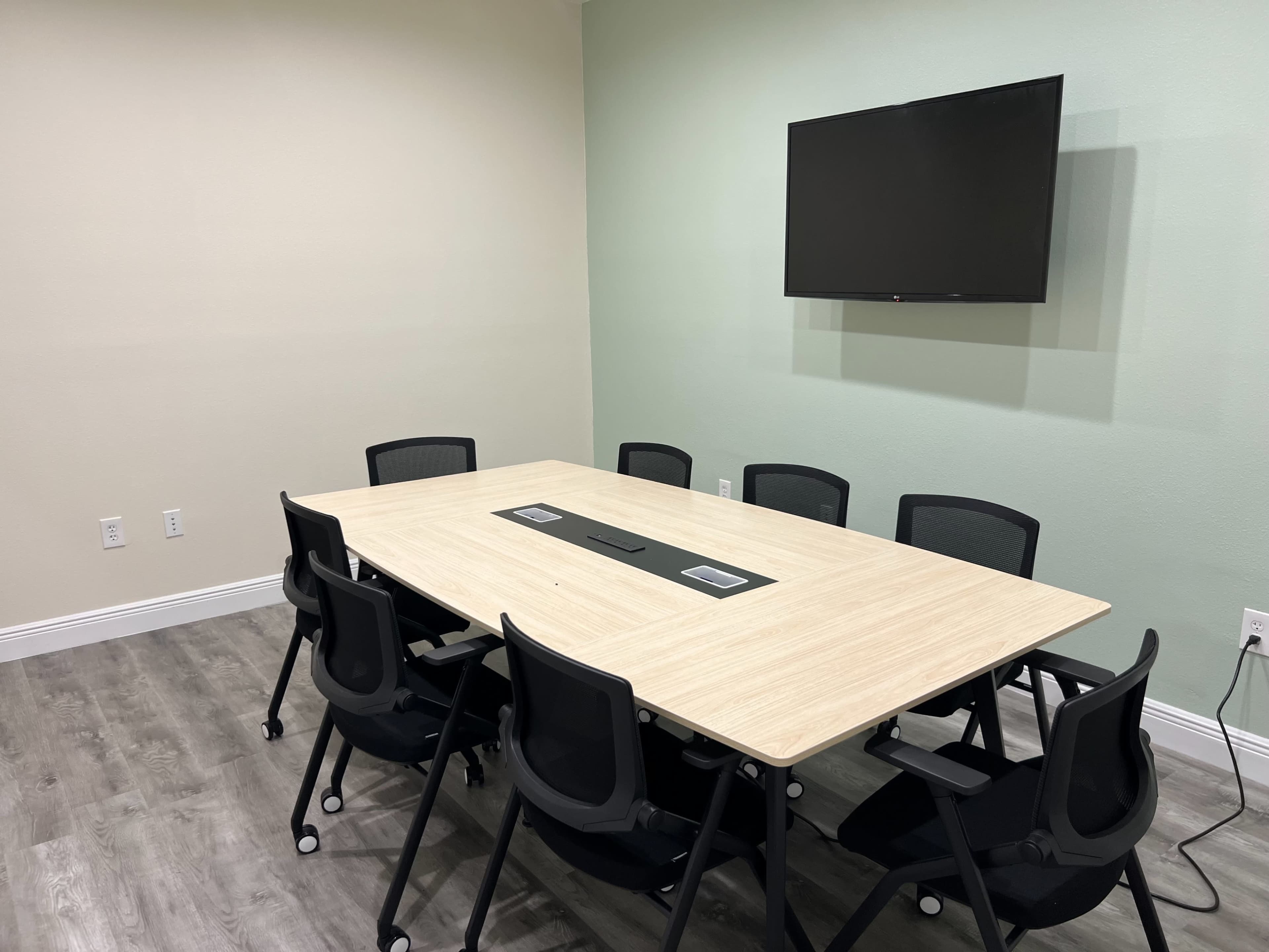 The image shows a conference room with a large wooden table surrounded by eight black chairs and a wall-mounted television.