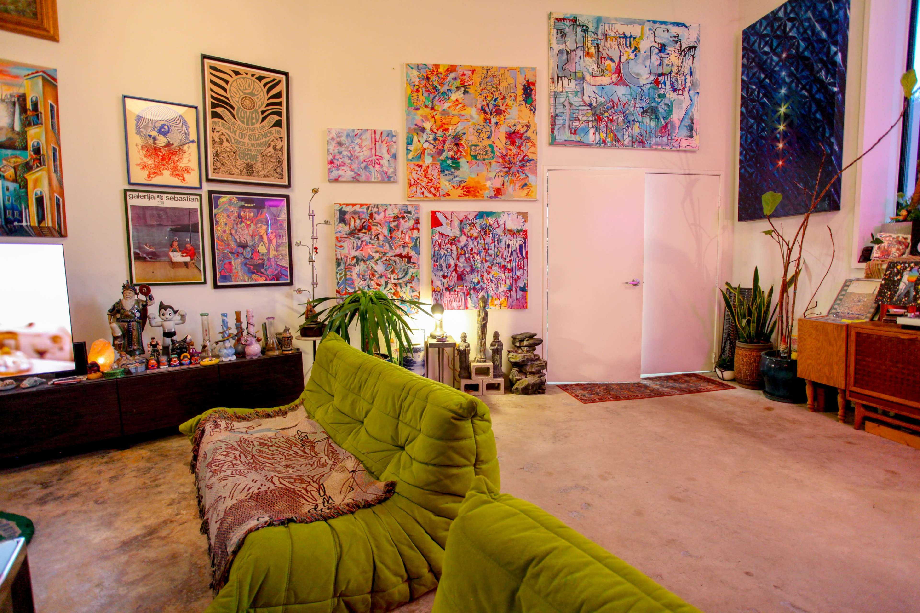 A colorful living space features a green sofa, various artworks on the walls, and indoor plants.