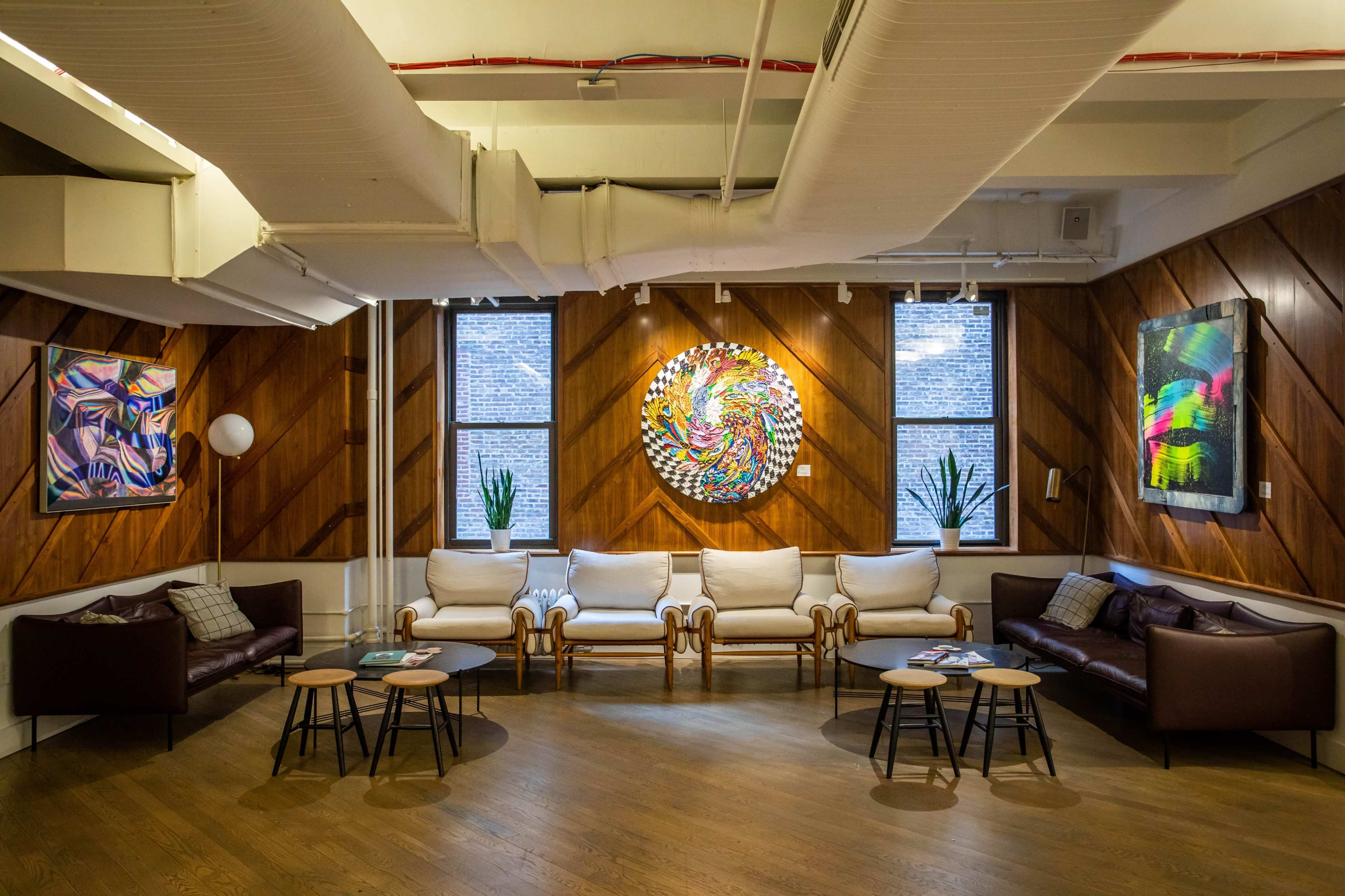 A modern lounge area with two brown sofas, a set of small round tables, and colorful abstract artwork displayed on the wooden walls.