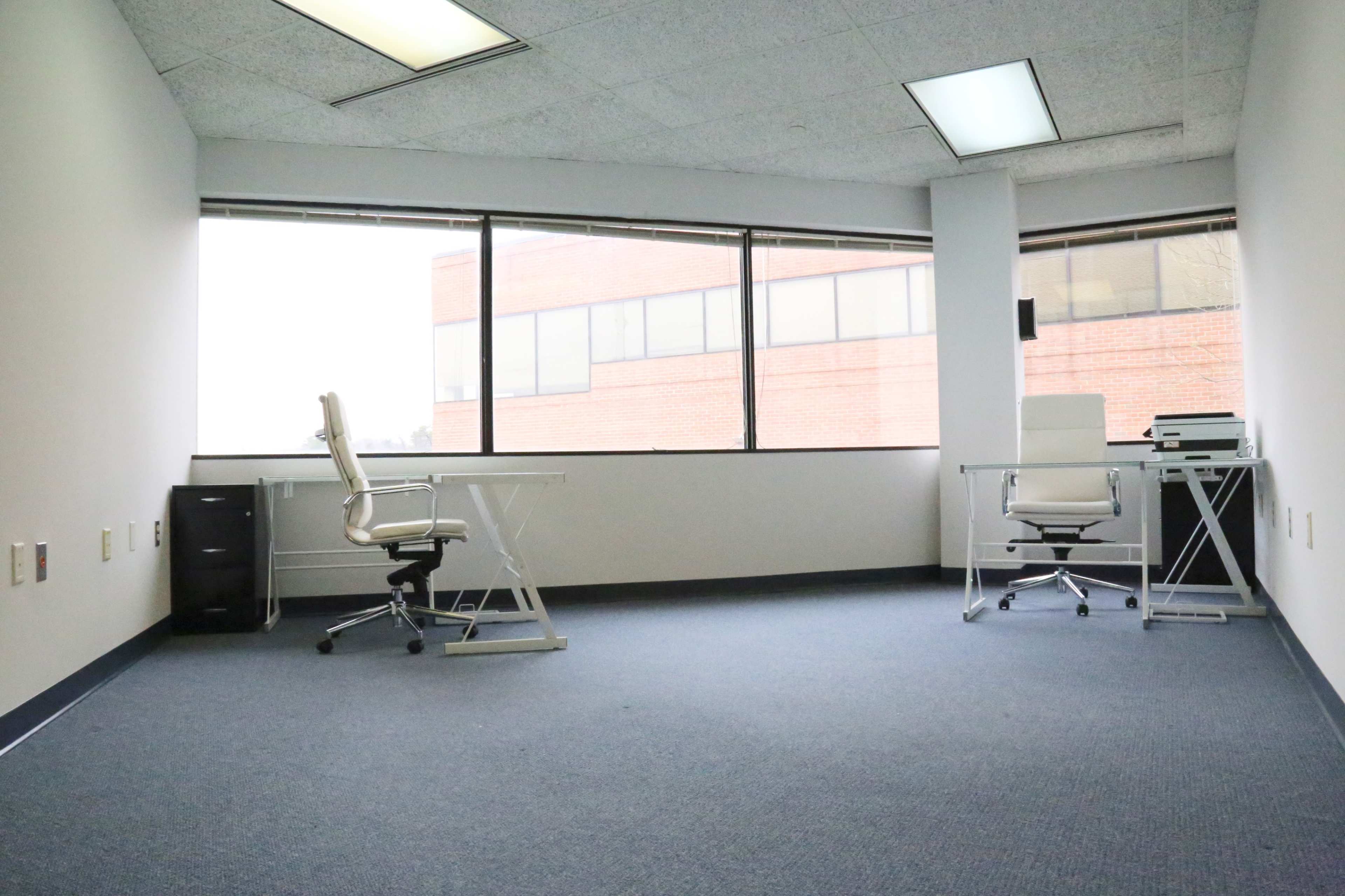 A vacant office space features two white desks with chairs and large windows overlooking a brick building.