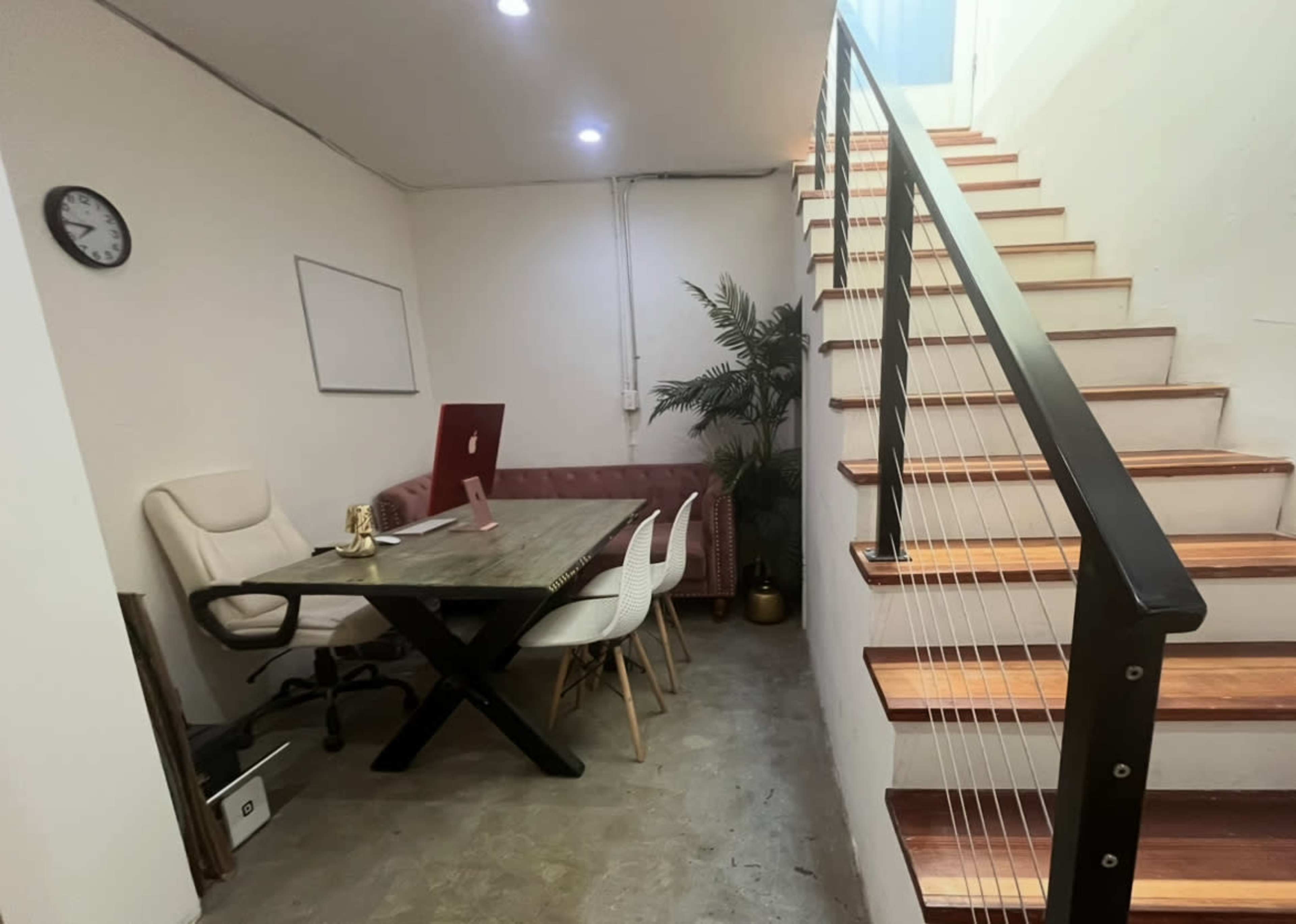 The image depicts a small office space featuring a desk with a computer, a whiteboard, a chair, and a small potted plant, alongside a staircase with wooden steps and a railing.