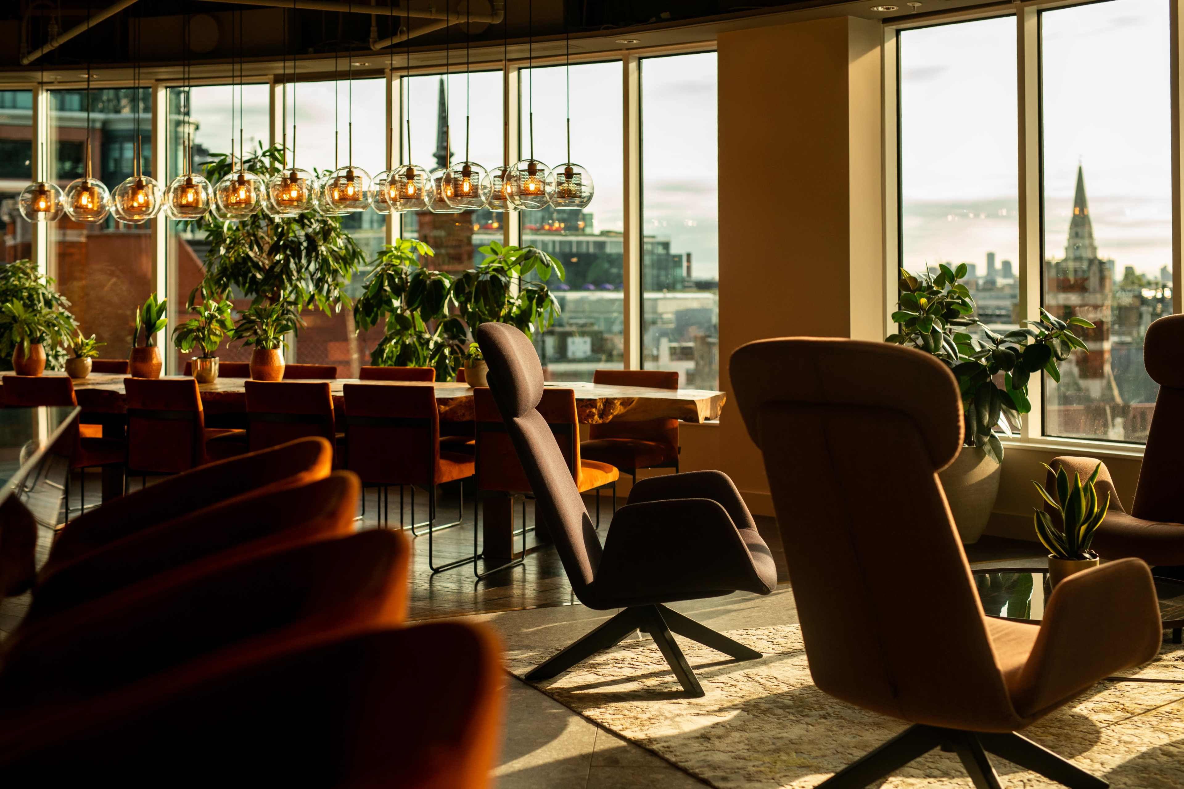 The image shows a modern dining area with large windows, several potted plants, and stylish furniture, including a long table and elegant chairs, set against a cityscape backdrop.