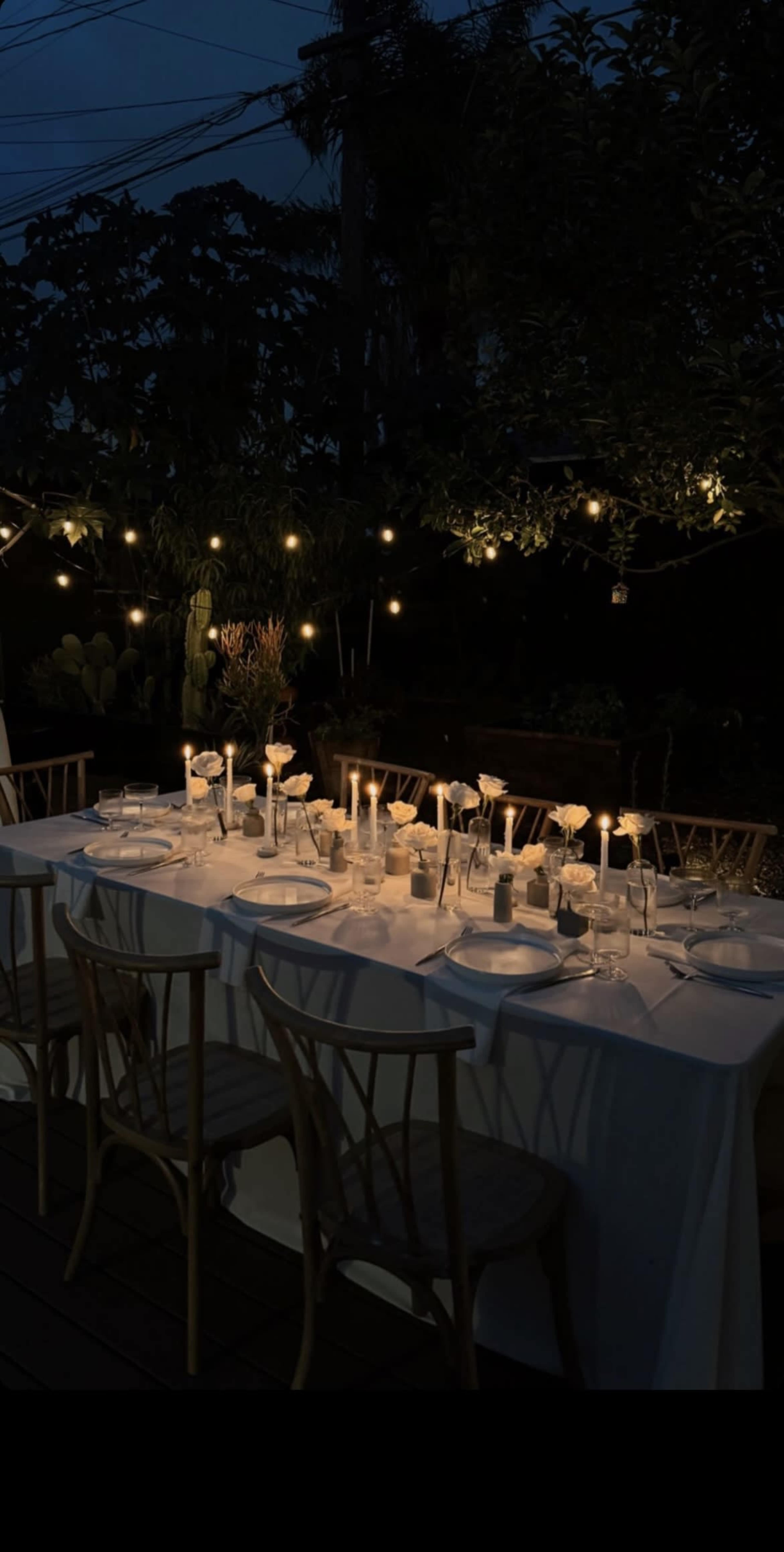 A long table is set for dinner outdoors at night, illuminated by candles and string lights, surrounded by greenery.