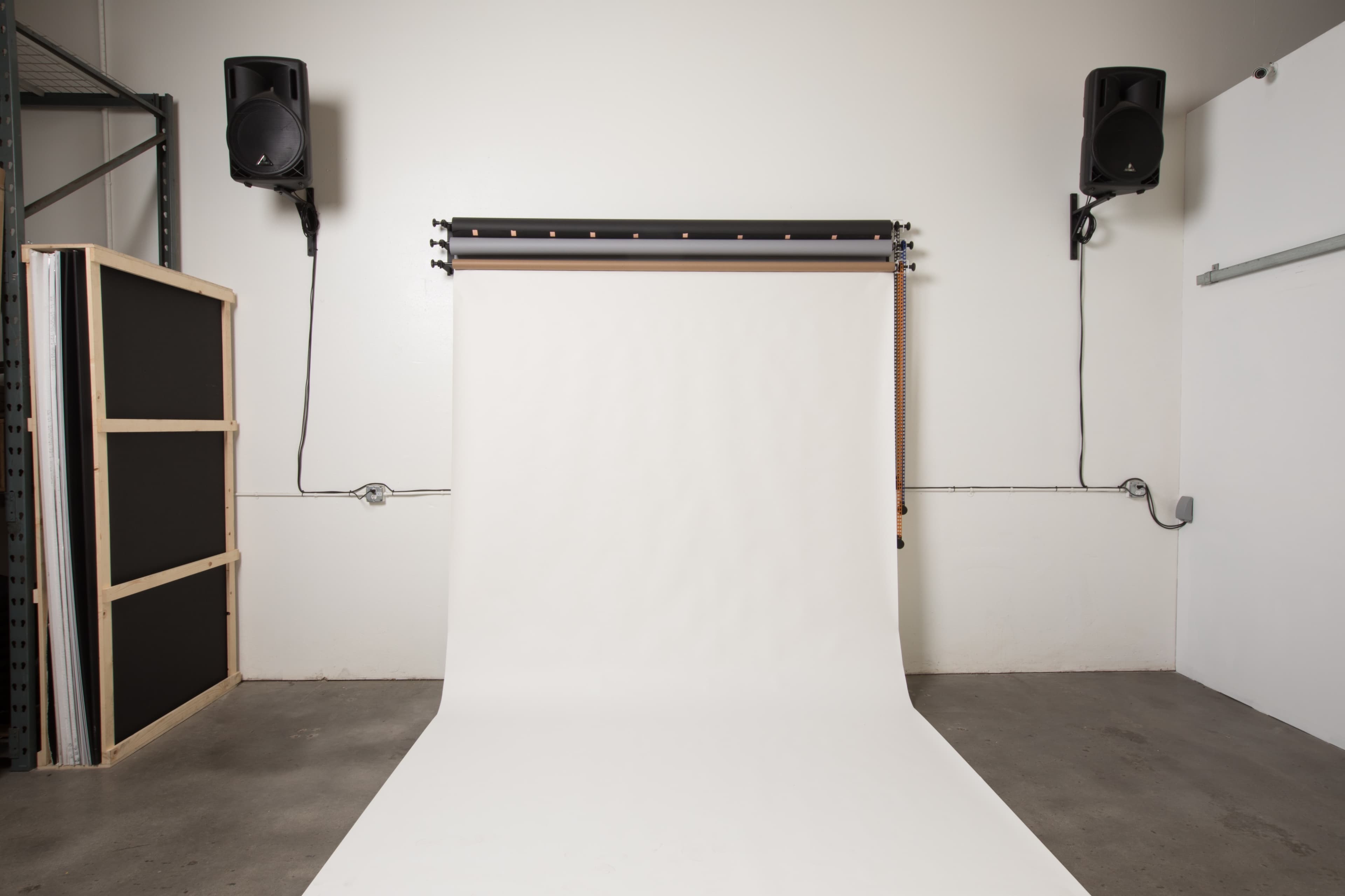 A photography studio with a plain white backdrop, flanked by two speakers mounted on the wall.