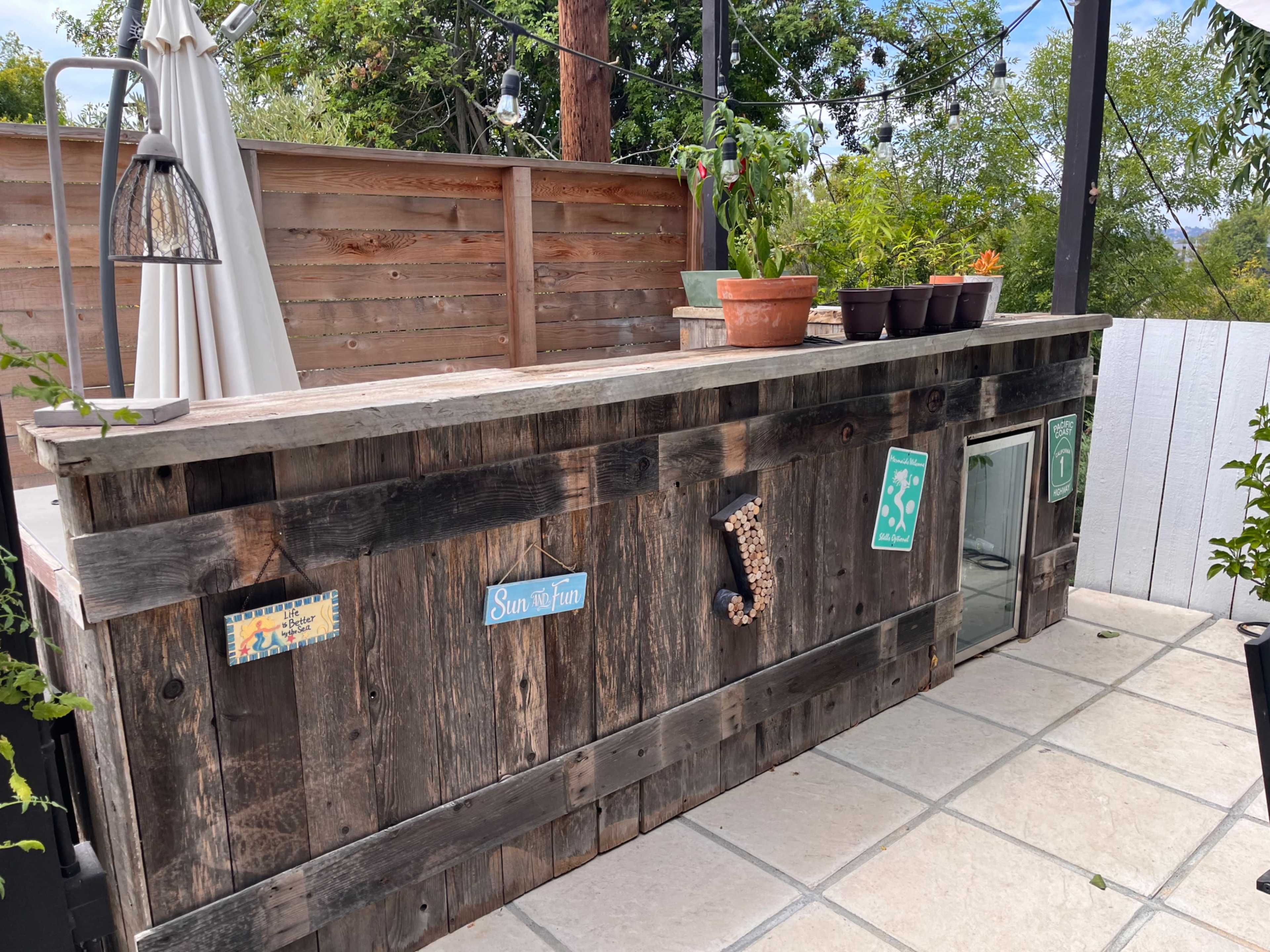 A weathered wooden bar with a countertop, adorned with potted plants and decorative signs, situated on a tiled patio surrounded by greenery.