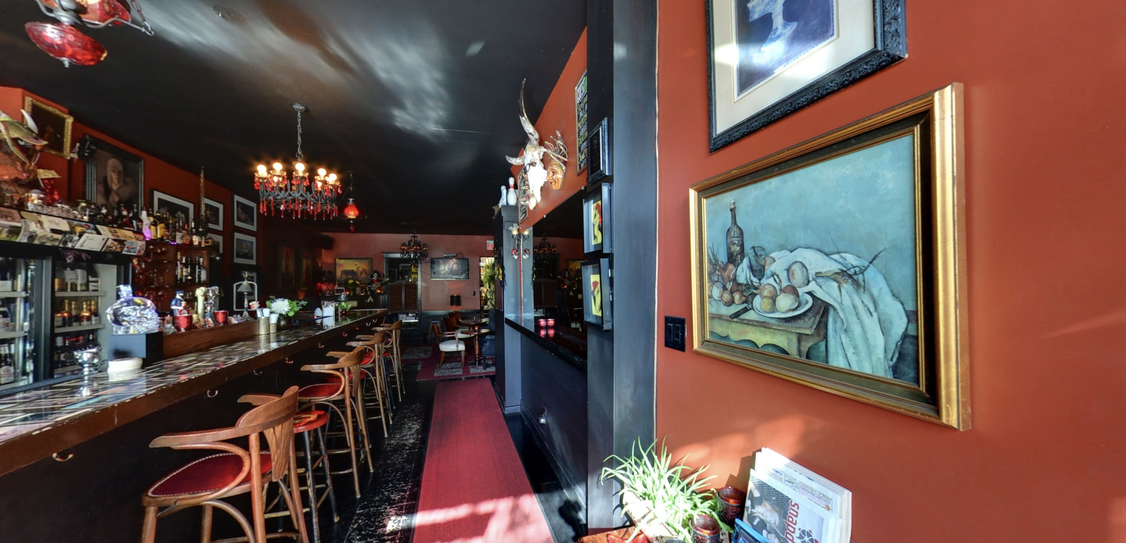 The image shows the interior of a bar with a dark color scheme, featuring a long wooden counter, bar stools, and a wall adorned with framed paintings and decorative items.