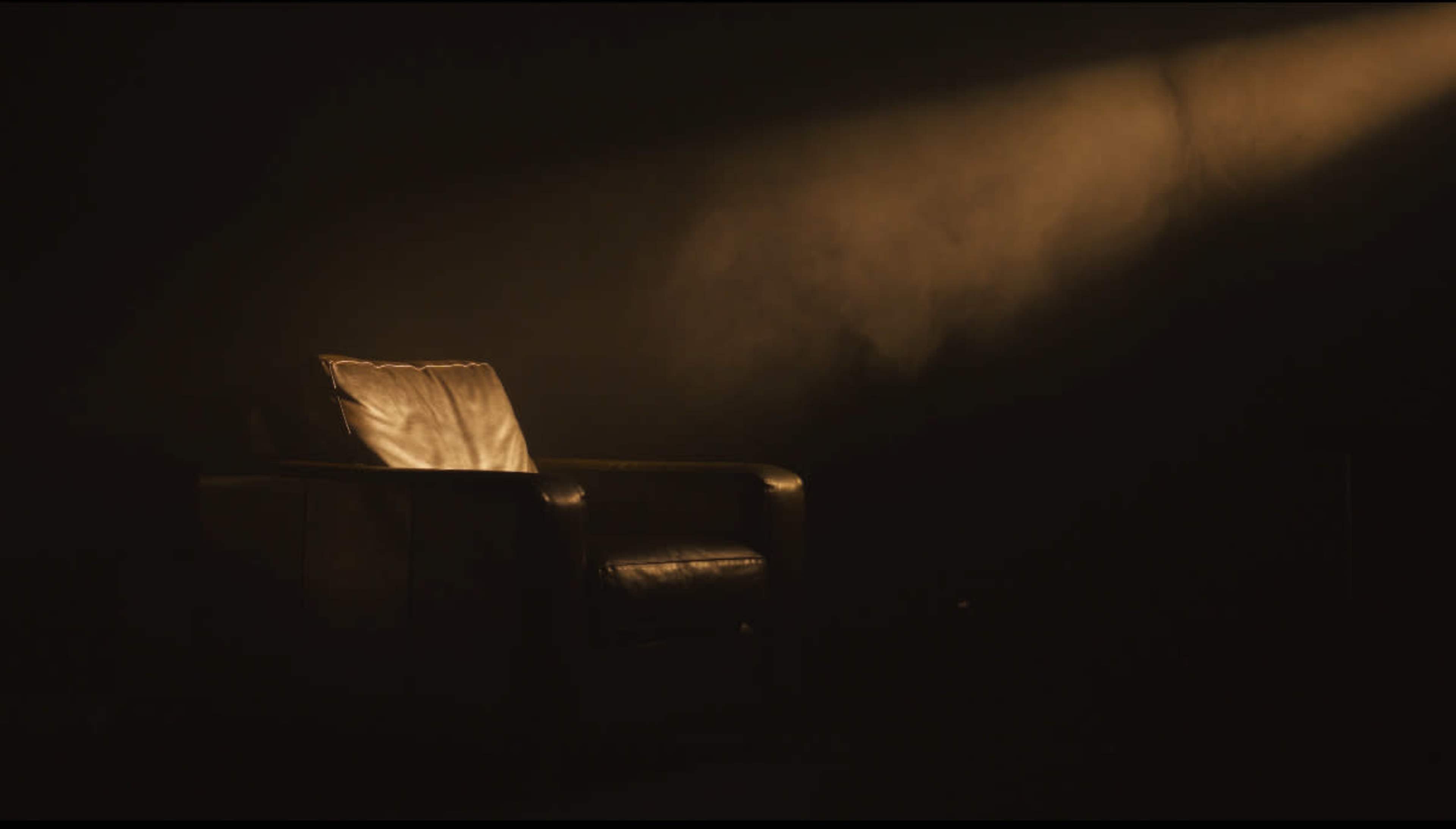 A single leather chair sits in a dimly lit space with a beam of light falling on it.