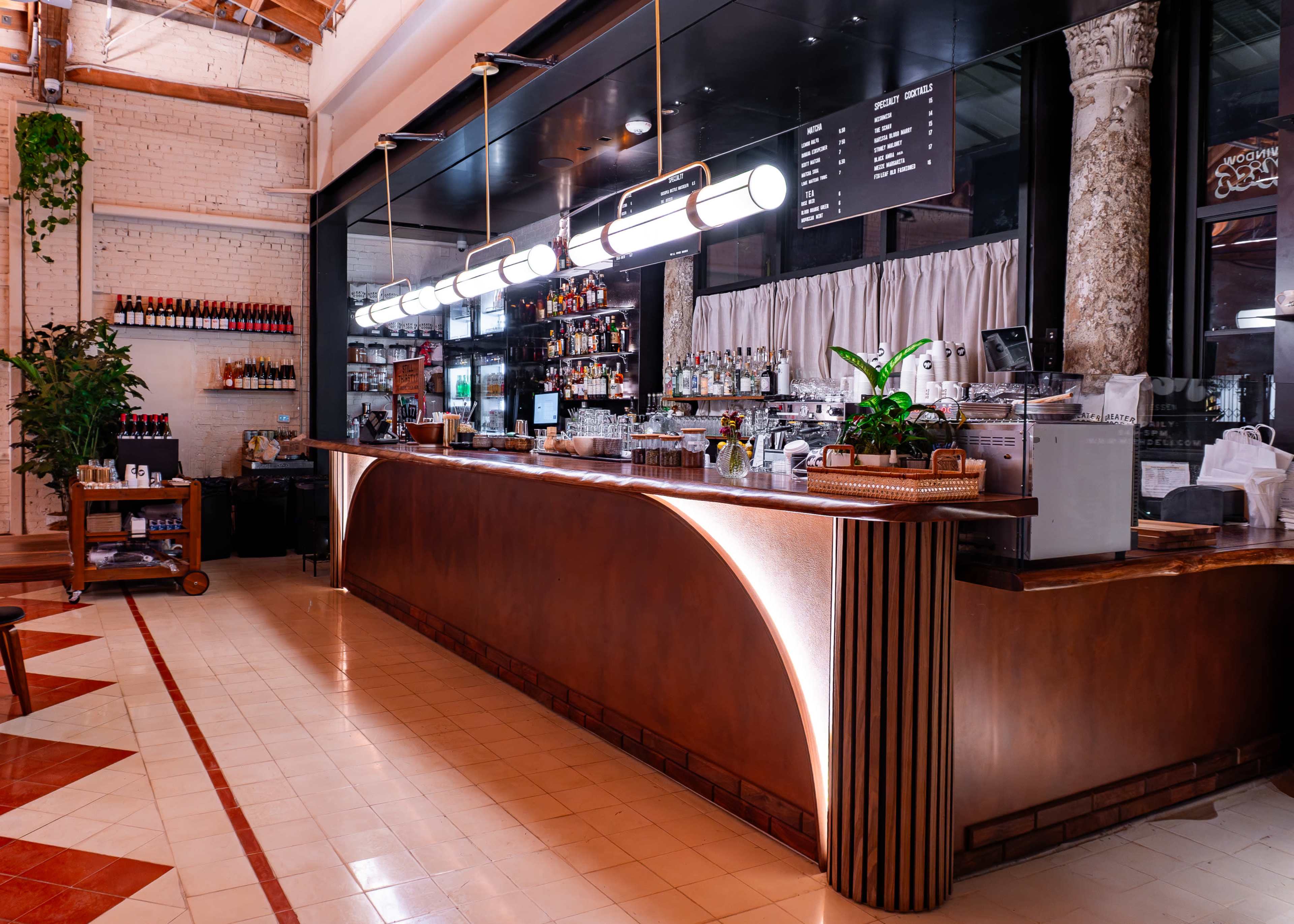 The image shows a modern café interior featuring a long wooden bar with bright overhead lighting, a variety of bottles on shelves, and a tiled floor with a geometric pattern.