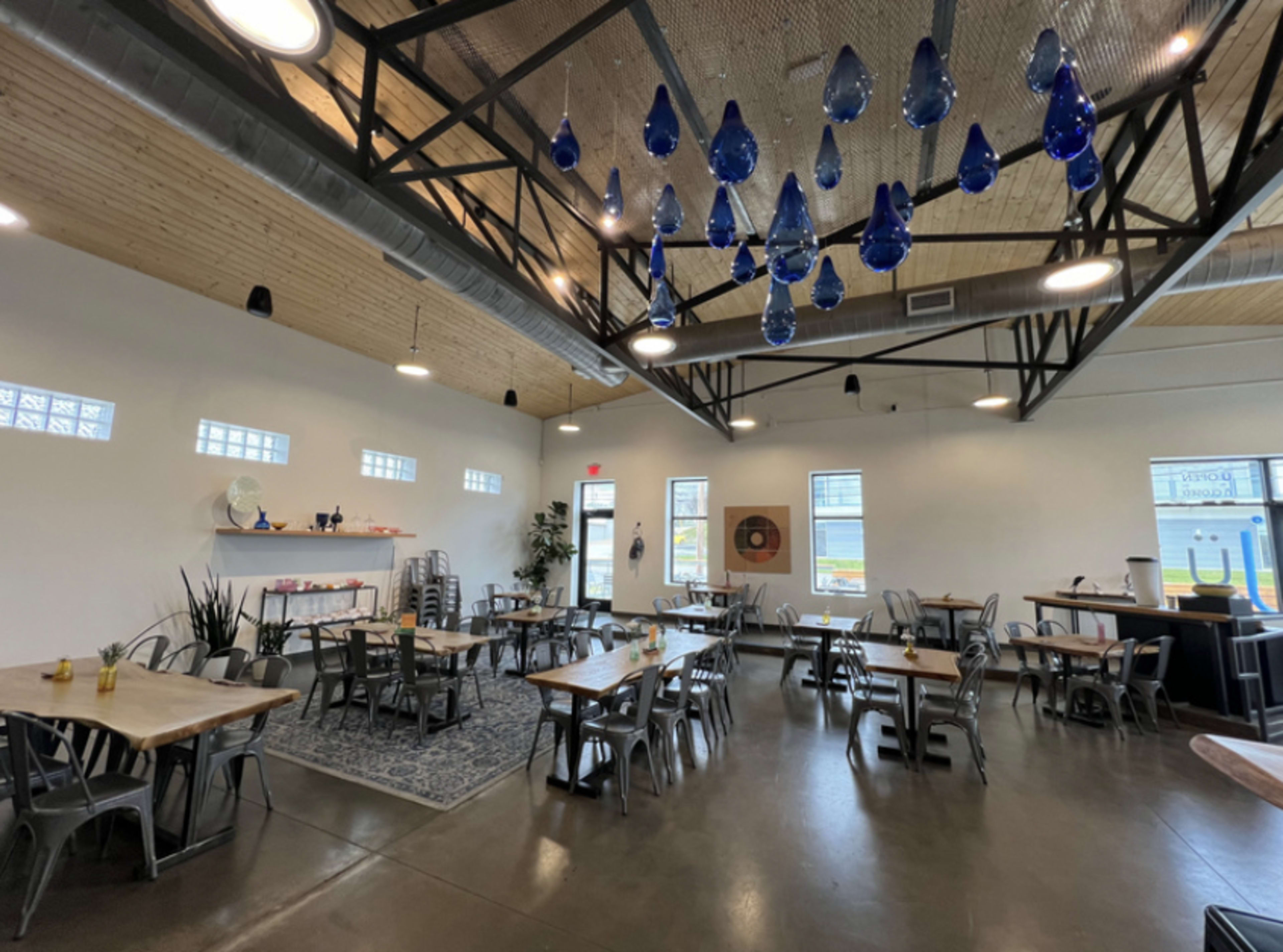 A modern restaurant interior with wooden tables, metal chairs, and blue glass pendants hanging from the ceiling.