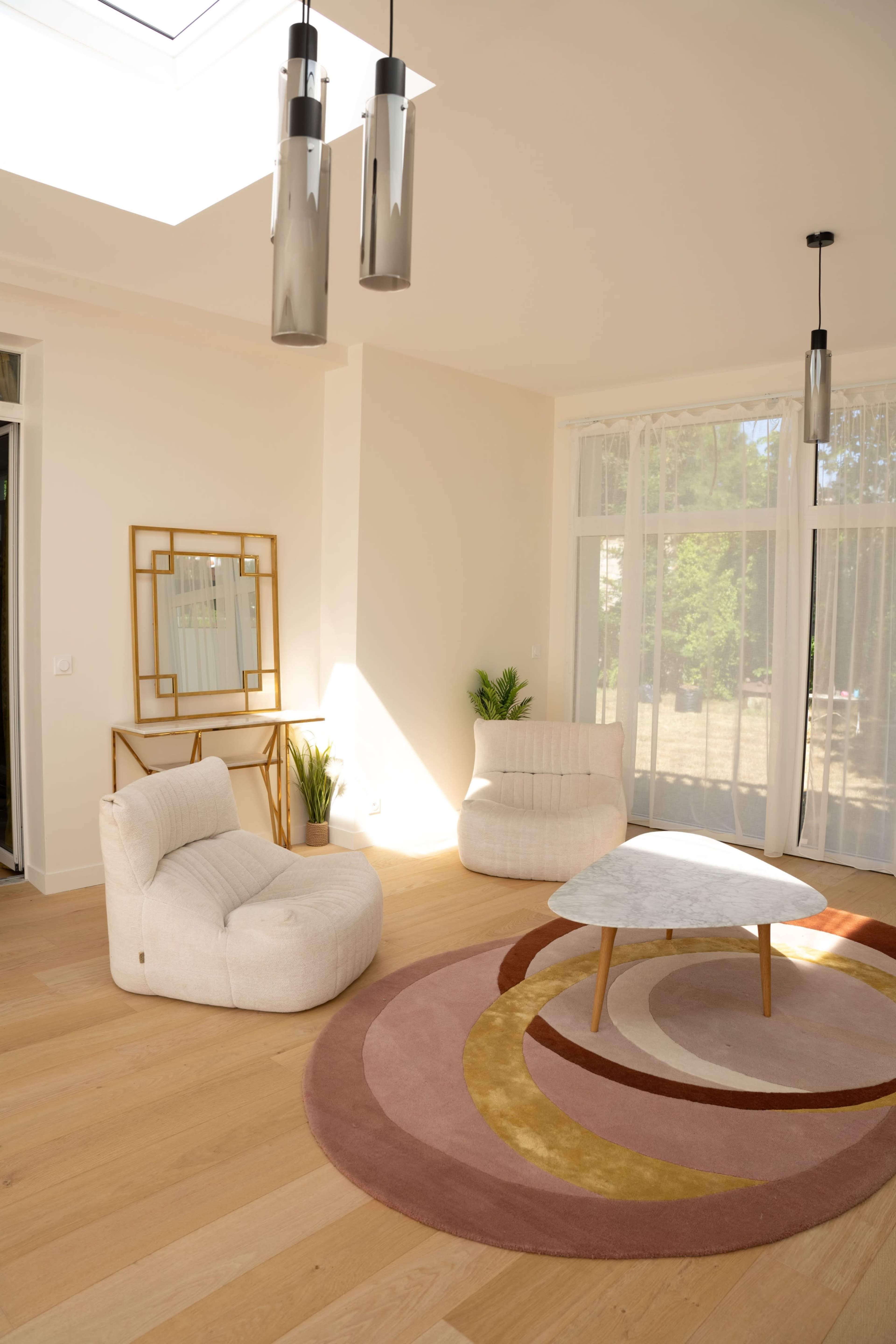The image shows a well-lit interior space featuring two plush armchairs, a circular area rug with concentric designs, and a modern table beneath pendant lights.