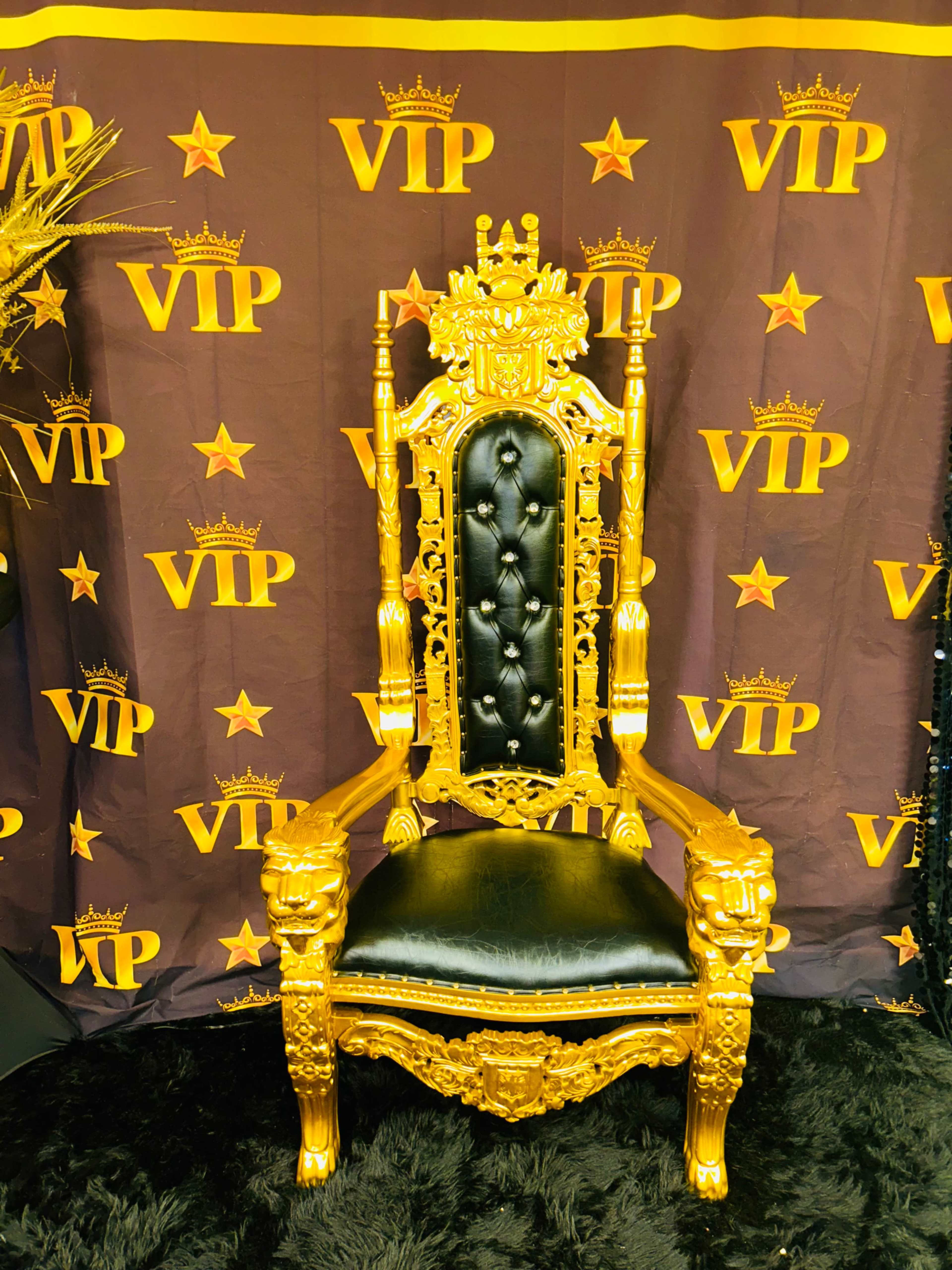 An ornate golden throne with a black padded seat, situated against a backdrop displaying the letters "VIP" and star motifs.
