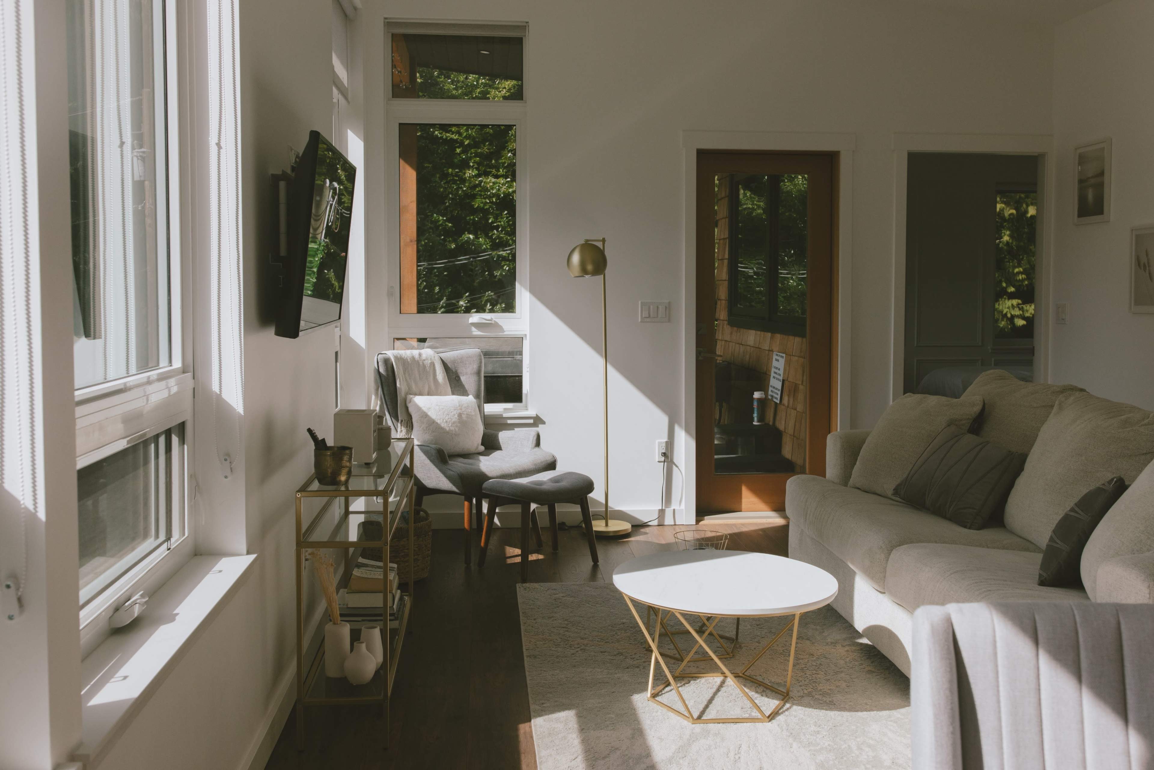 A bright living room features a gray sofa, a gold-accented coffee table, and a television mounted on one wall, with large windows allowing natural light to illuminate the space.