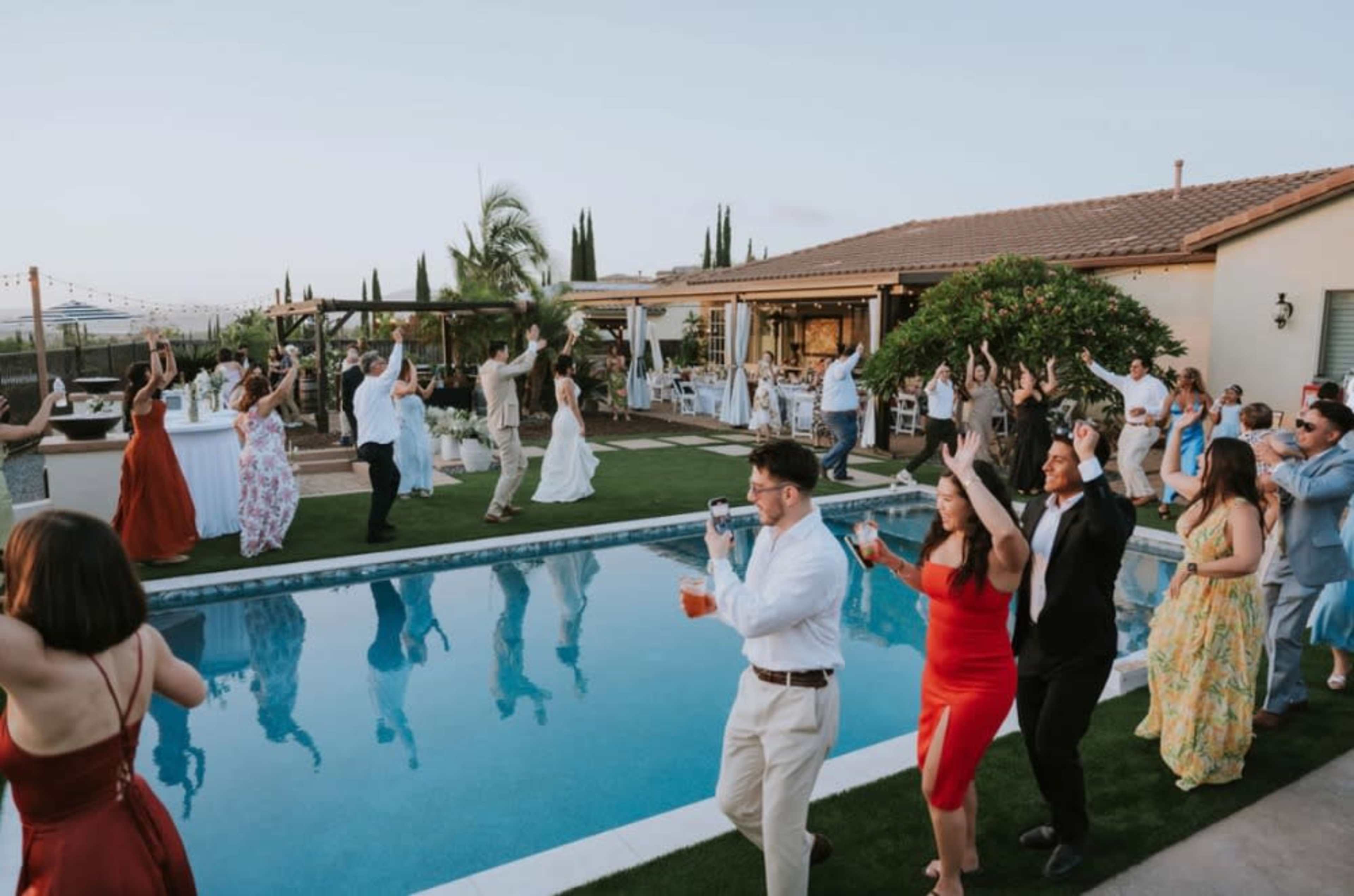 A lively outdoor evening scene features guests dancing by a pool at a celebration, with tables set up nearby under a clear sky.
