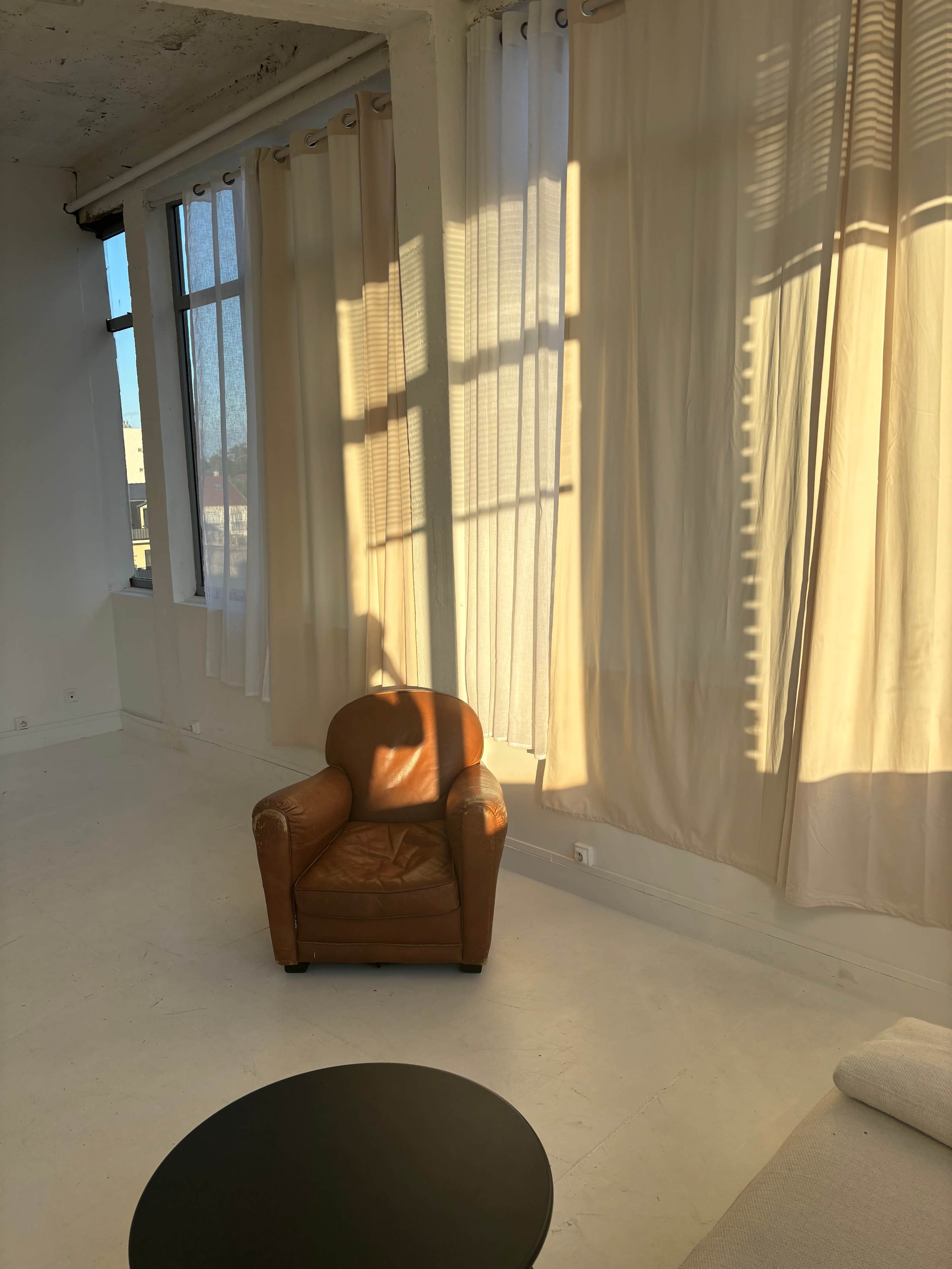 A brown leather chair sits beside a black circular table, with sunlight streaming through large windows draped in sheer curtains.