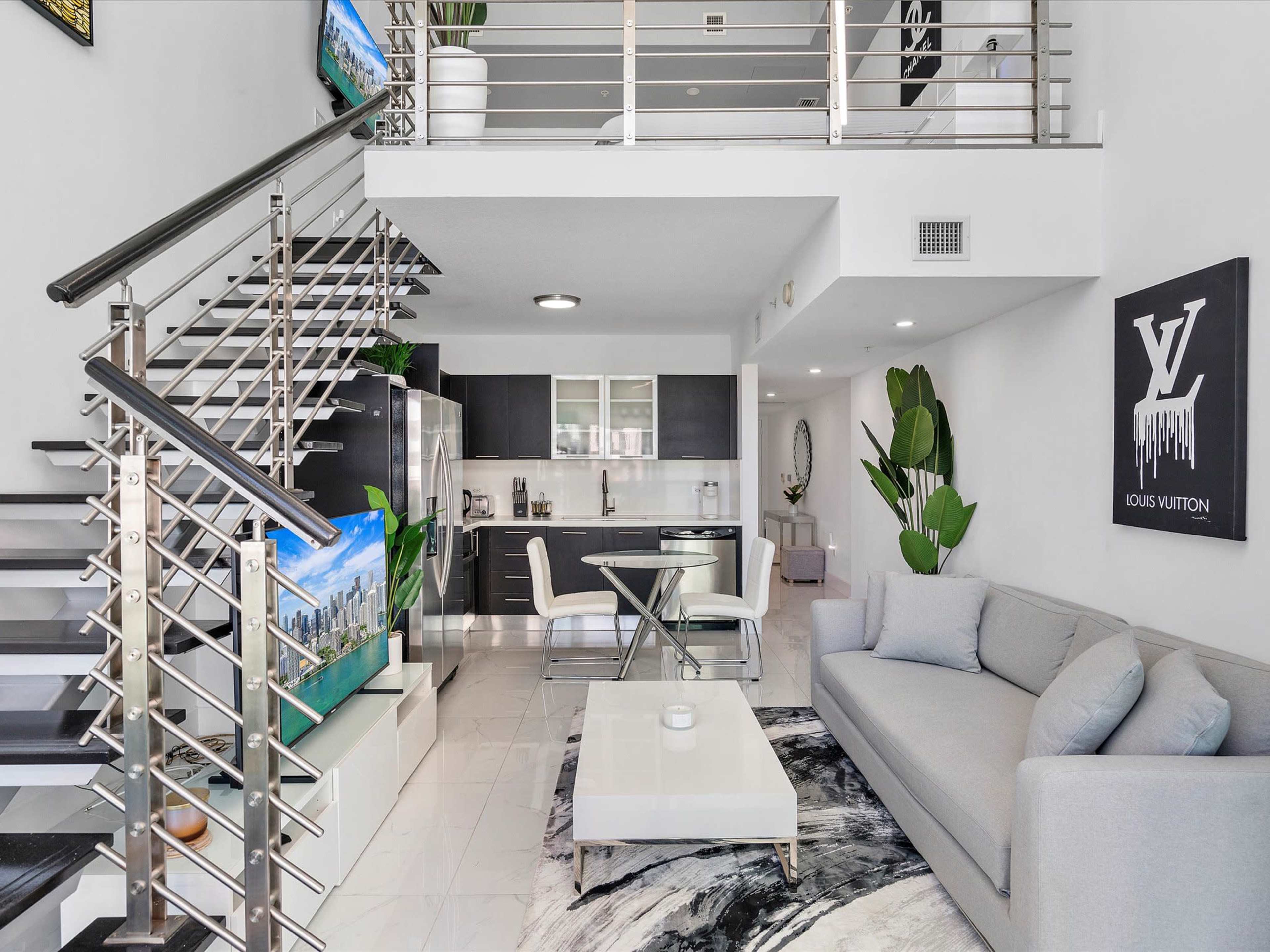 A modern living space featuring a gray sectional sofa, a glass dining table, and a staircase leading to a loft area, with contemporary decor and appliances.