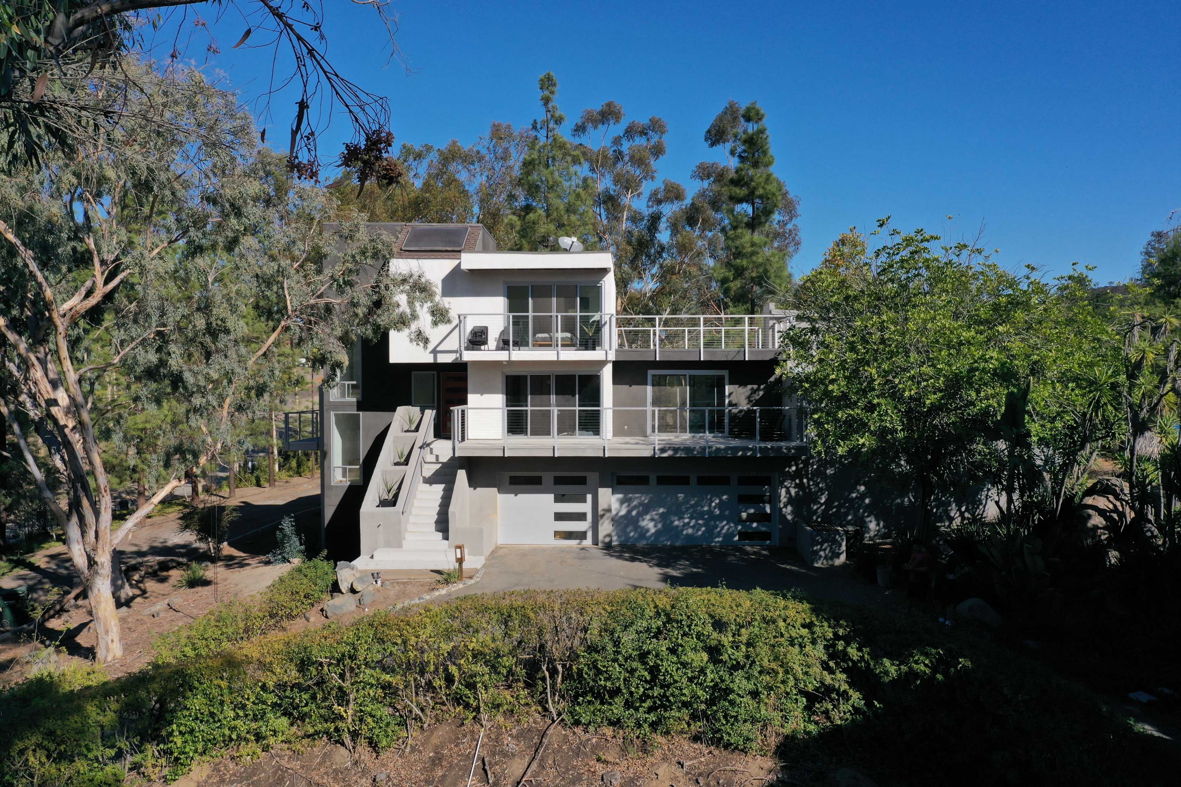 A modern two-story house with a flat roof is situated amidst trees, featuring a large front terrace and a driveway leading to a garage.