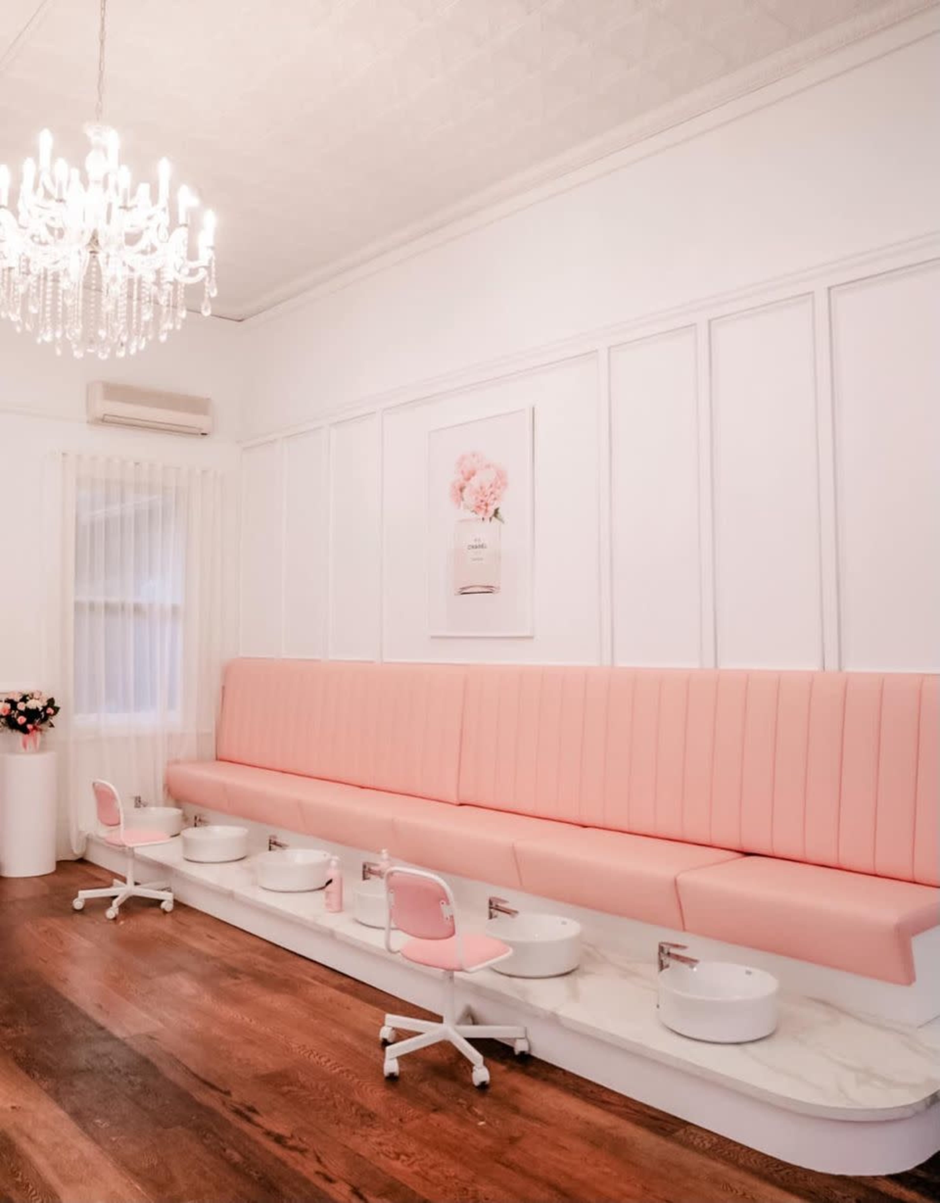 The image shows a brightly lit salon interior featuring a long pink bench with white pedicure stations and a crystal chandelier overhead.
