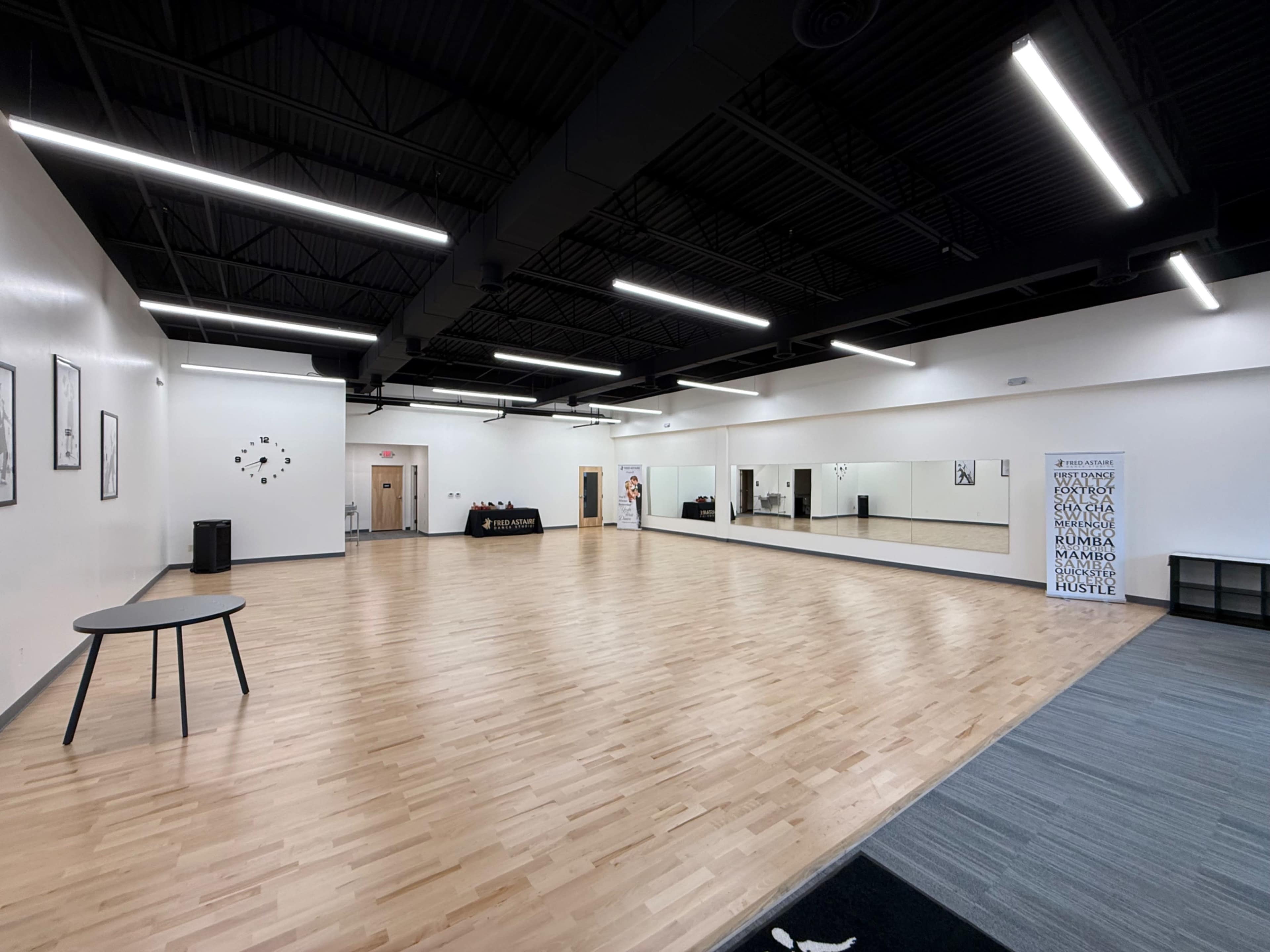 The image shows a spacious dance studio with a polished wooden floor, large mirrors on one wall, and a few pieces of furniture along the perimeter.