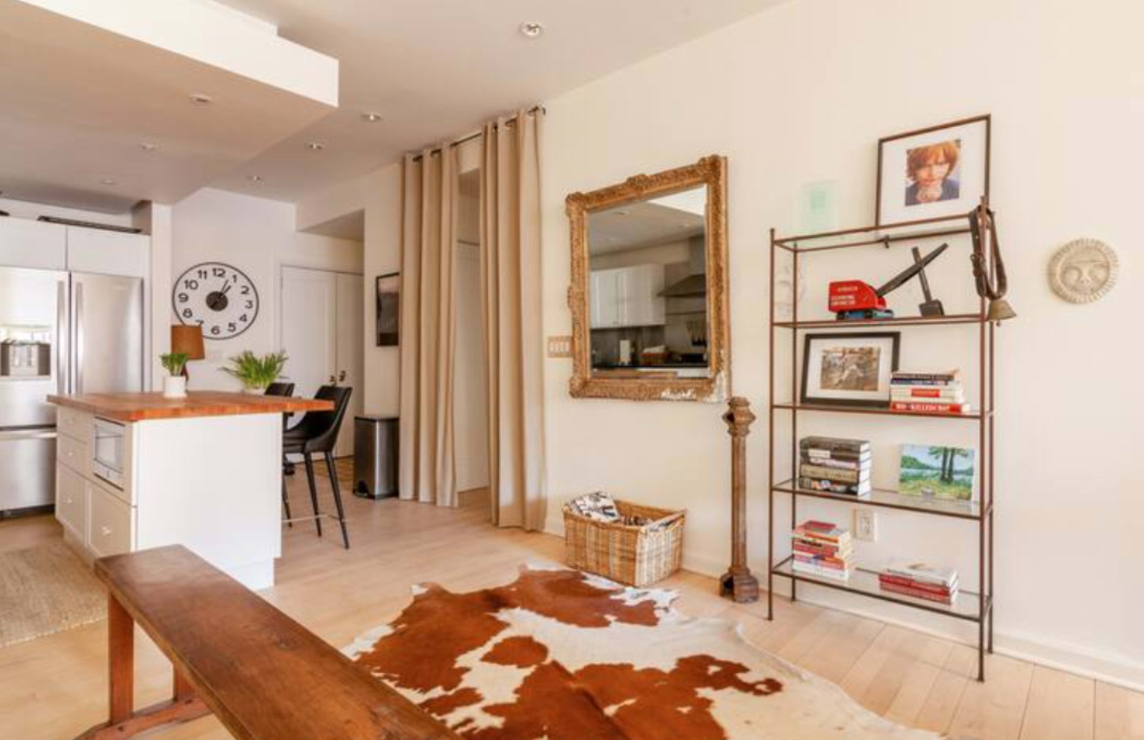 A modern living space with a wooden bench, a large mirror, a bookshelf filled with various items, and a kitchen area visible in the background.