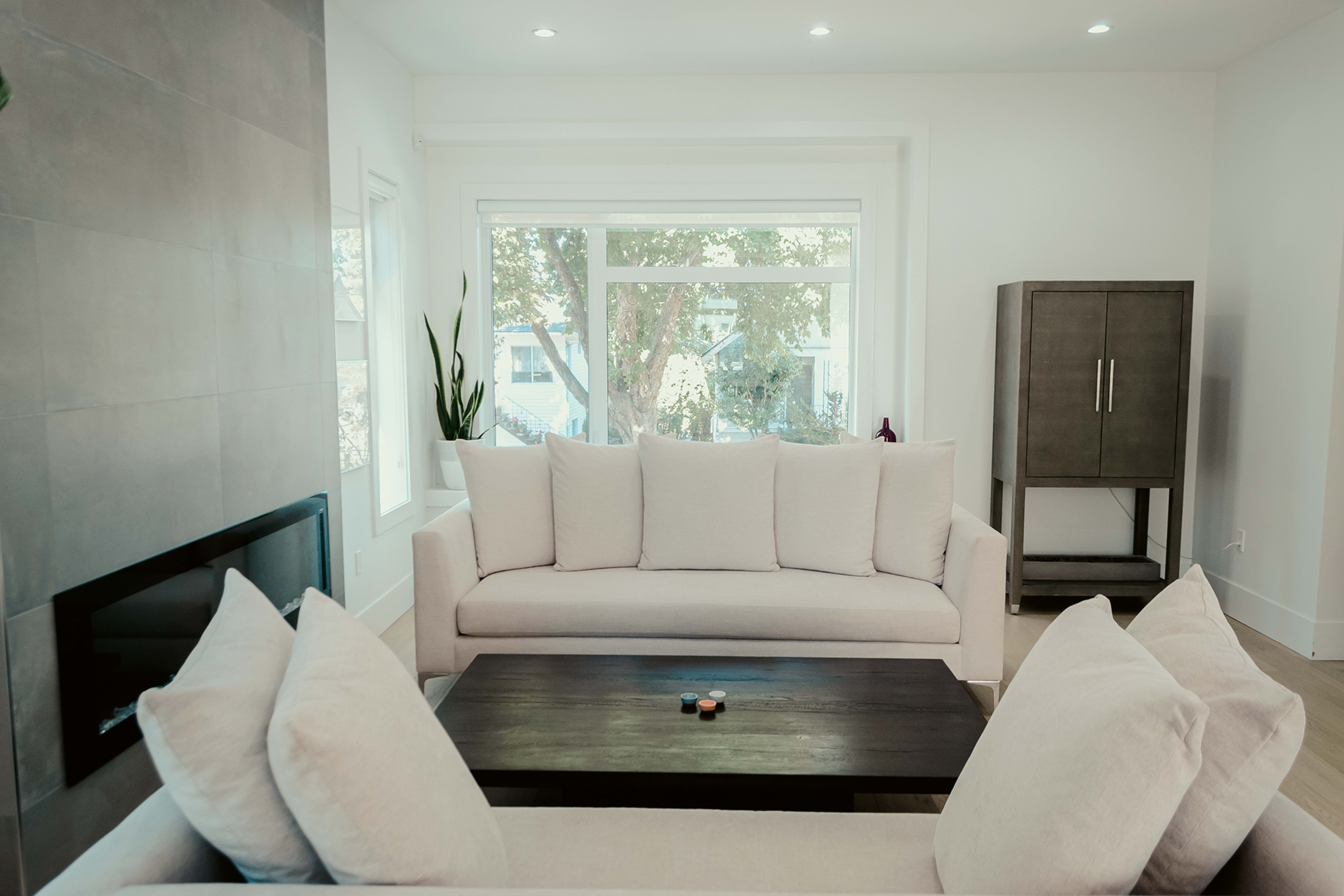 A bright living room features two light-colored sofas and a dark wooden coffee table, with large windows allowing natural light to fill the space.