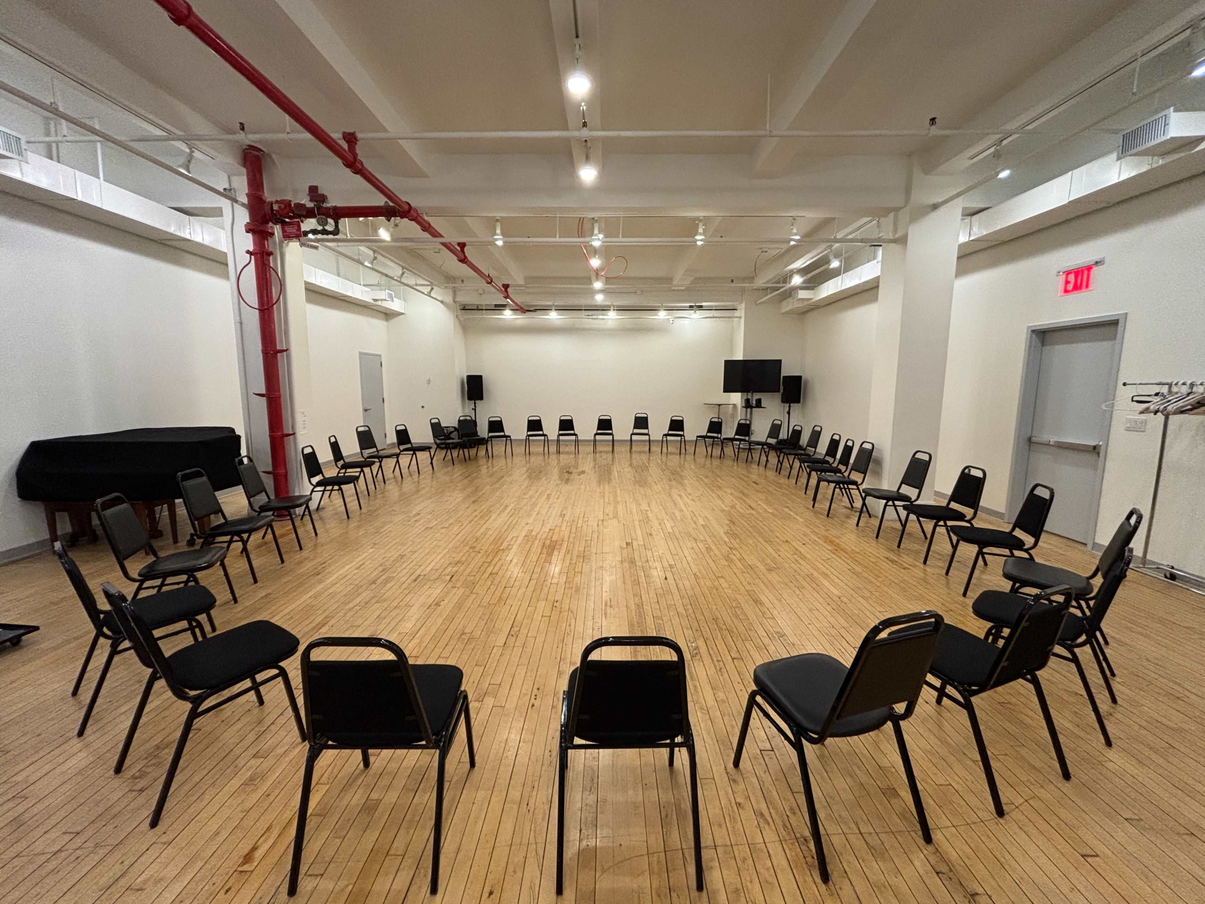 The image shows a large, empty room with wooden floors arranged in a circle of black chairs, facing inward.